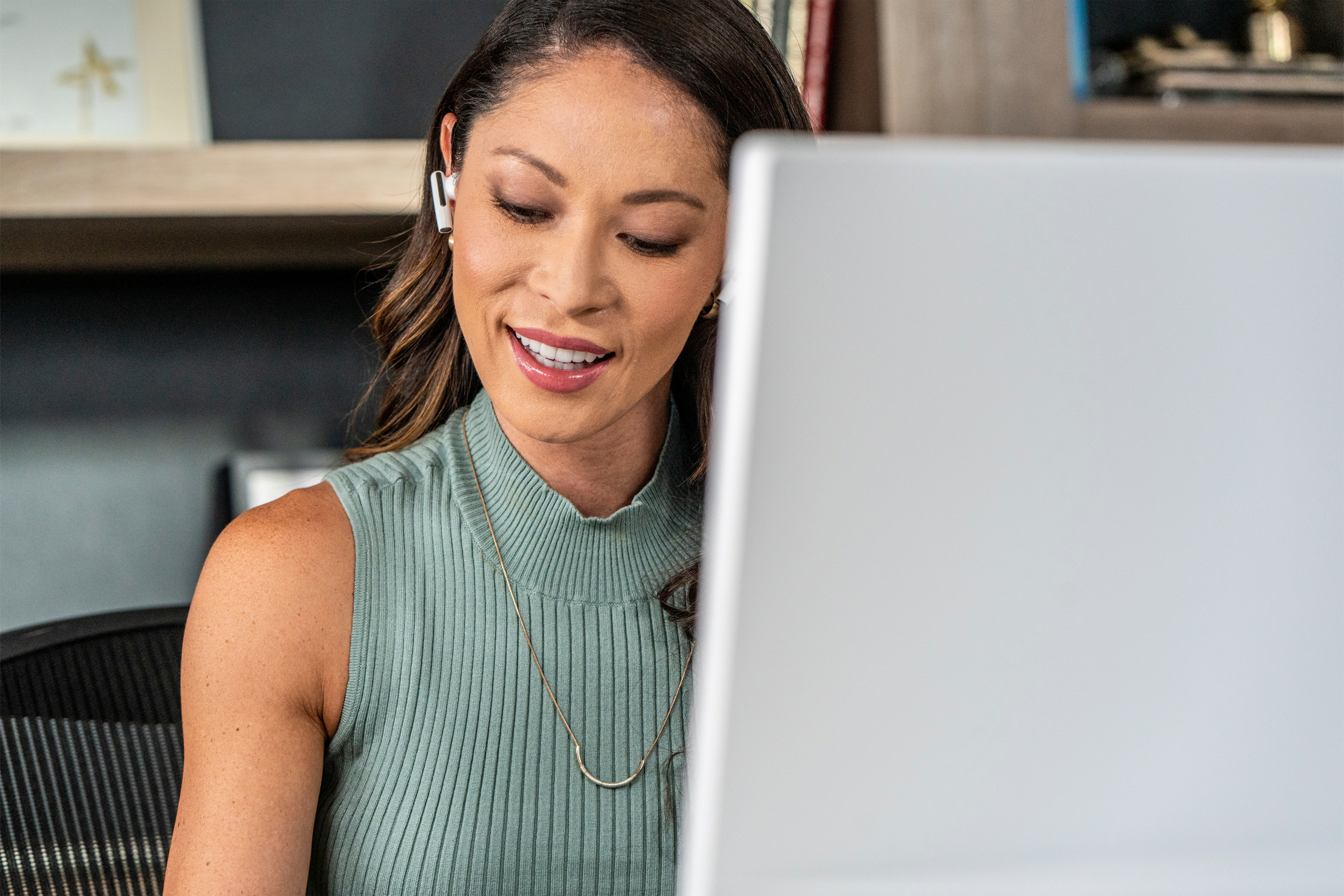Eine Frau lächelt, während sie auf einen Laptop-Bildschirm schaut, und ein Headset trägt. Eine Halskette ist sichtbar.