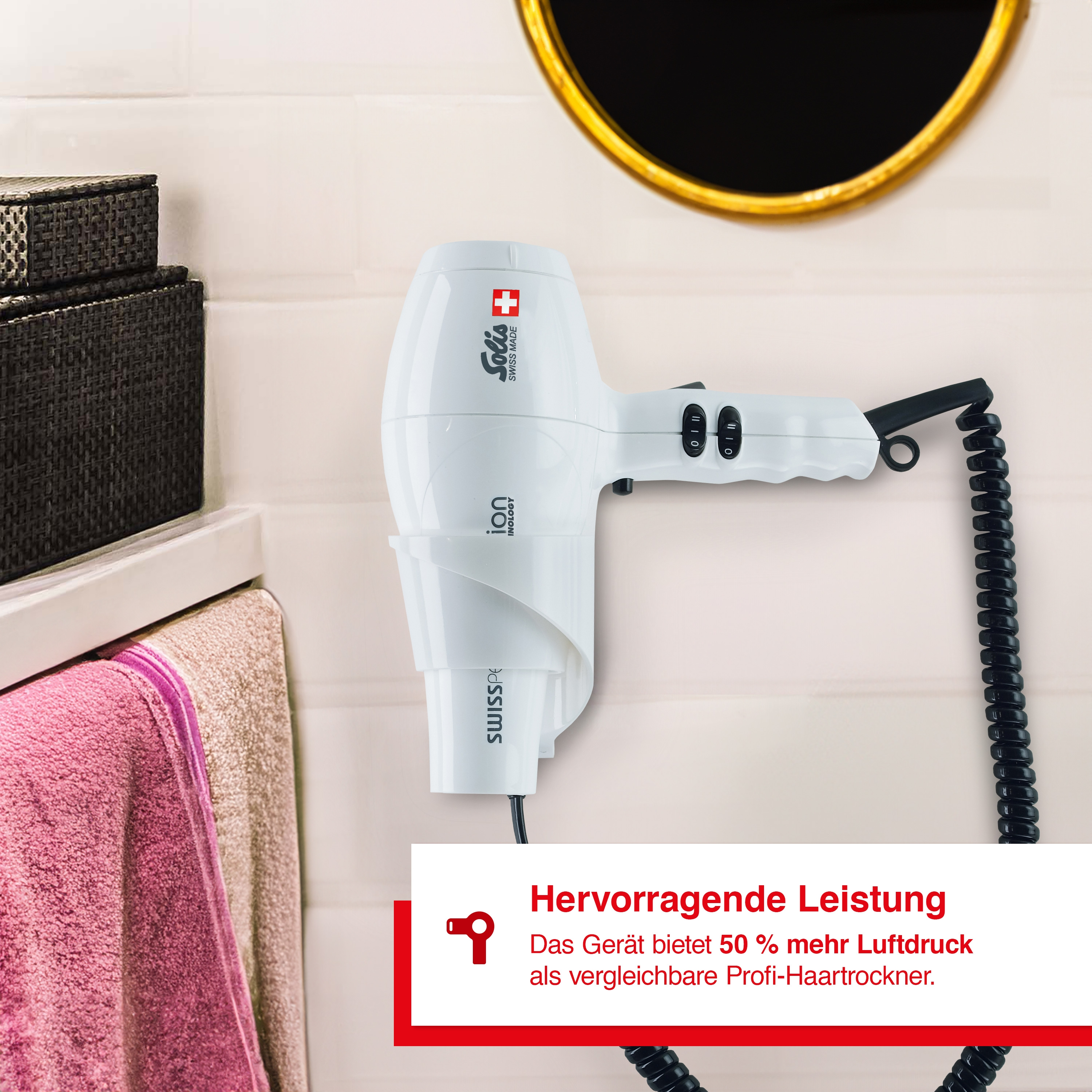 Ein weißer Haartrockner hängt an einer Wand, mit Spiegel und Handtuchhalter im Hintergrund.