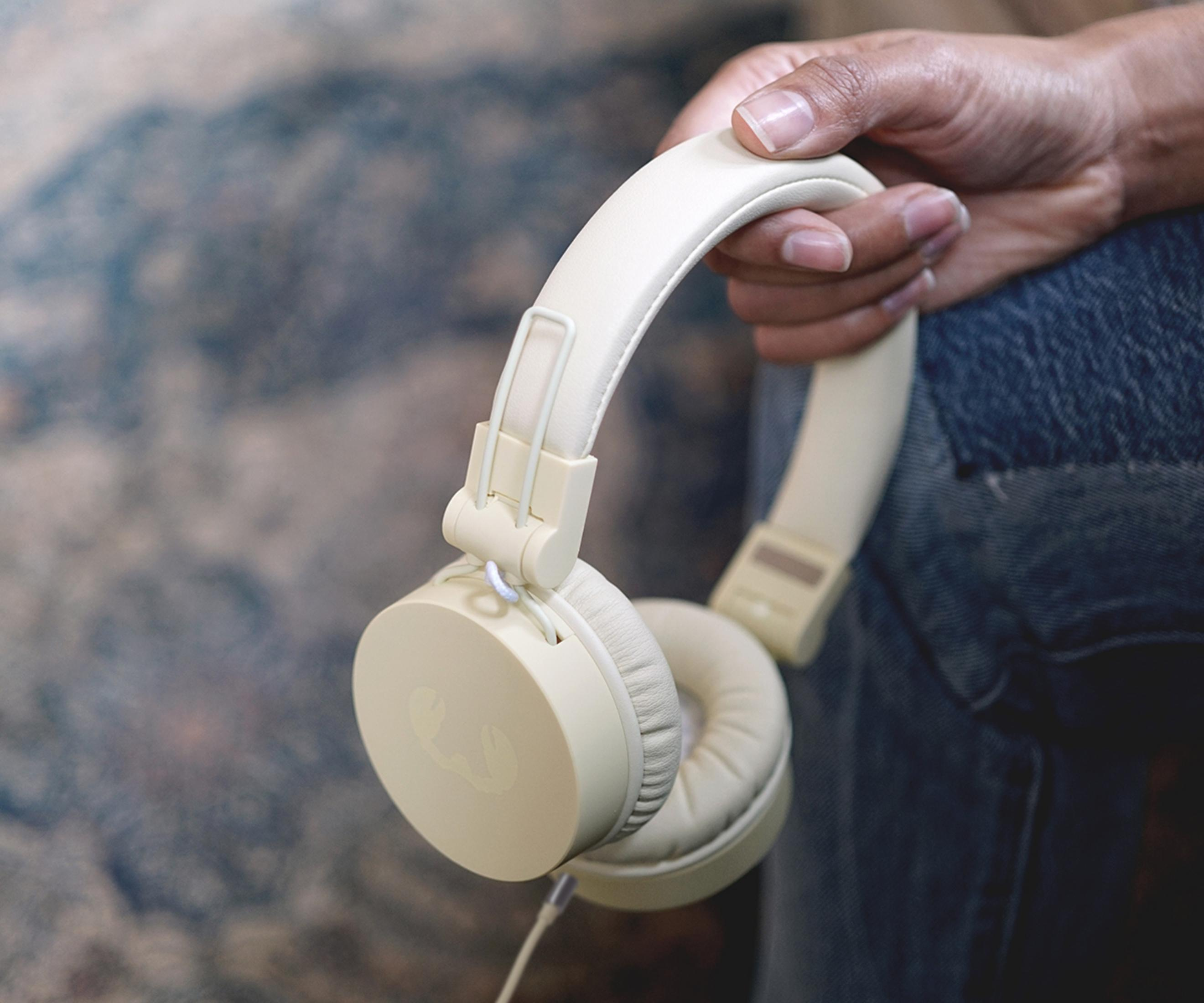 Osoba trzyma kremowe słuchawki. Są nauszne i mają biały kabel.