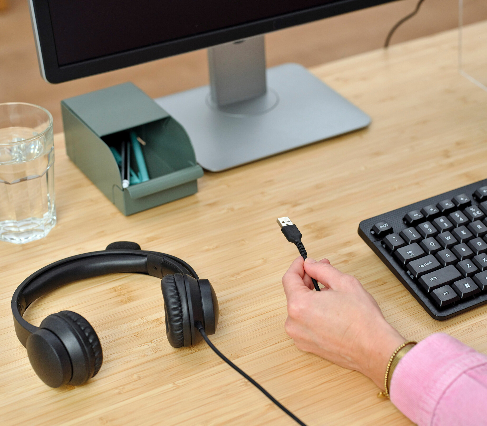 Ręce osoby w pobliżu biurka ze słuchawkami, monitorem i klawiaturą, trzymające kabel USB.