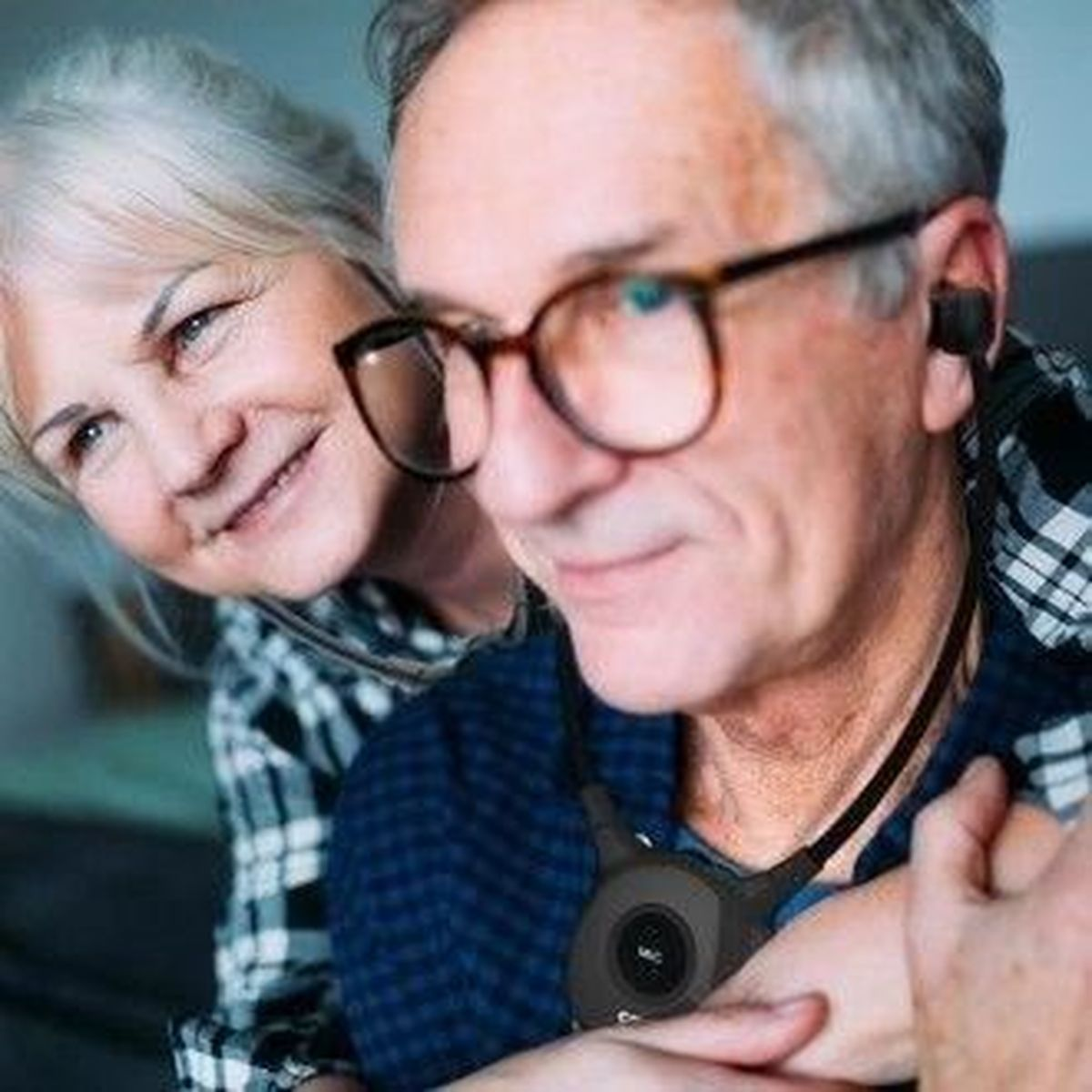 Coppia anziana, l'uomo ha auricolari e la donna sorride dietro di lui.