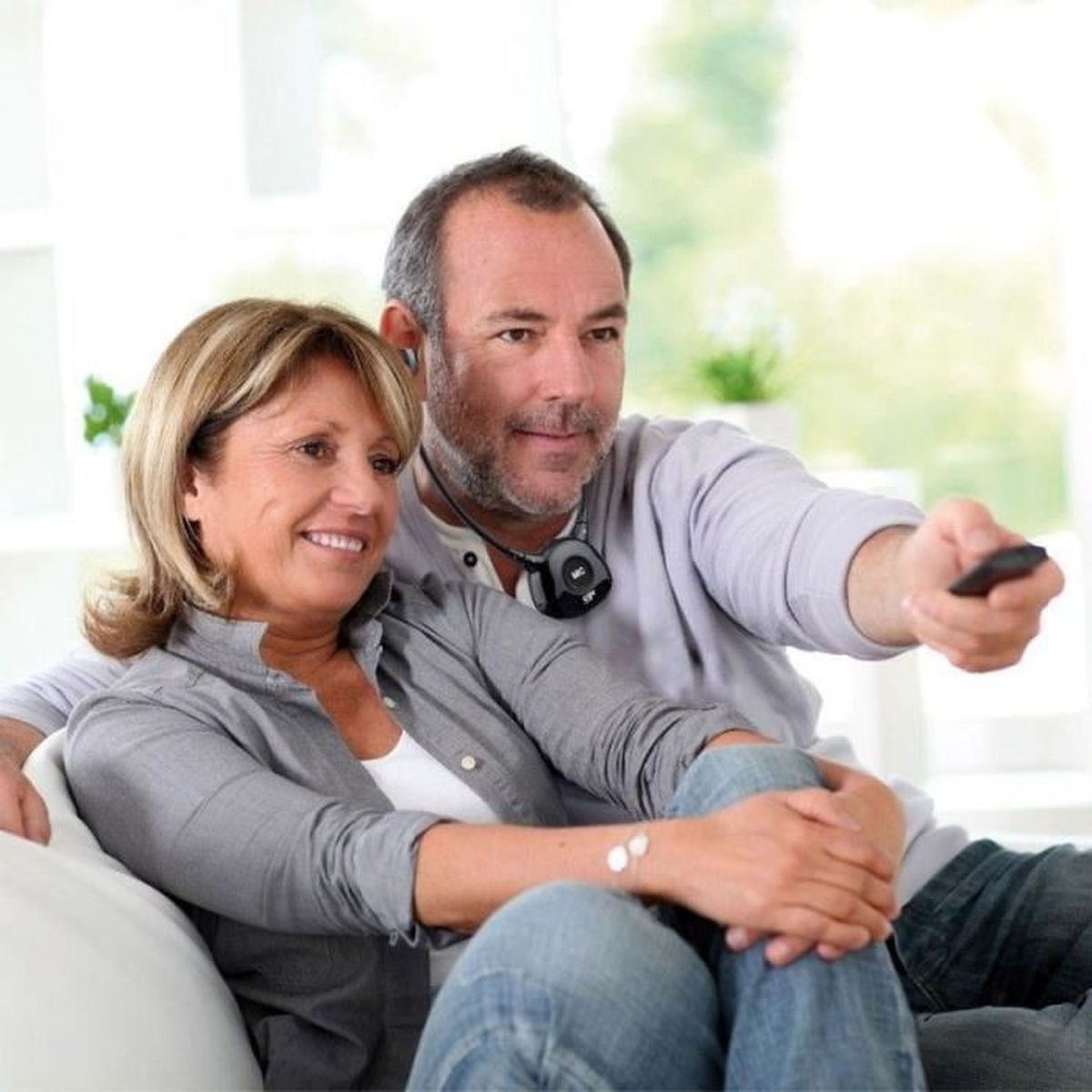 Una coppia sorridente seduta su un divano, che guarda la TV, con un amplificatore audio intorno al collo dell'uomo.