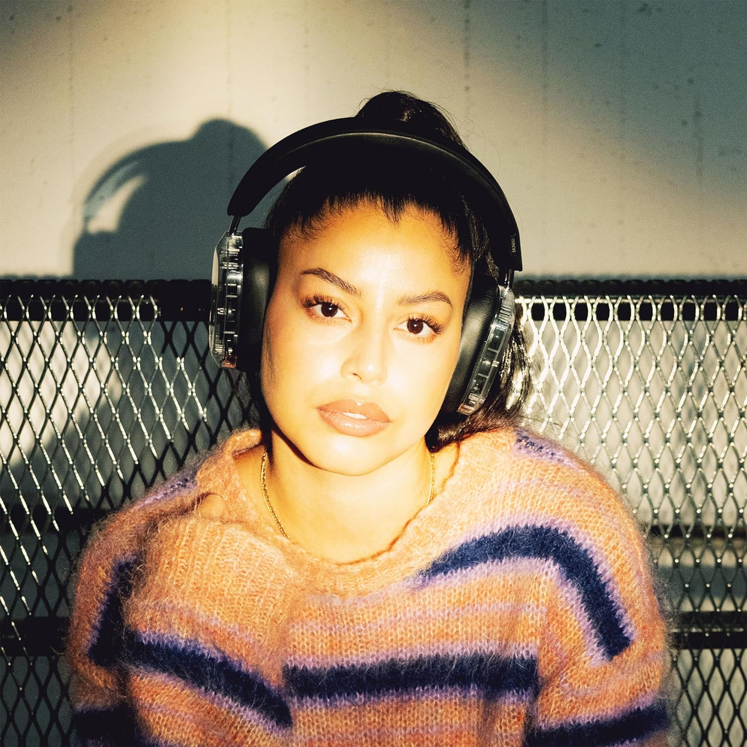 Kobieta w słuchawkach, w pasiastym swetrze. Tło ma metalową kratę.