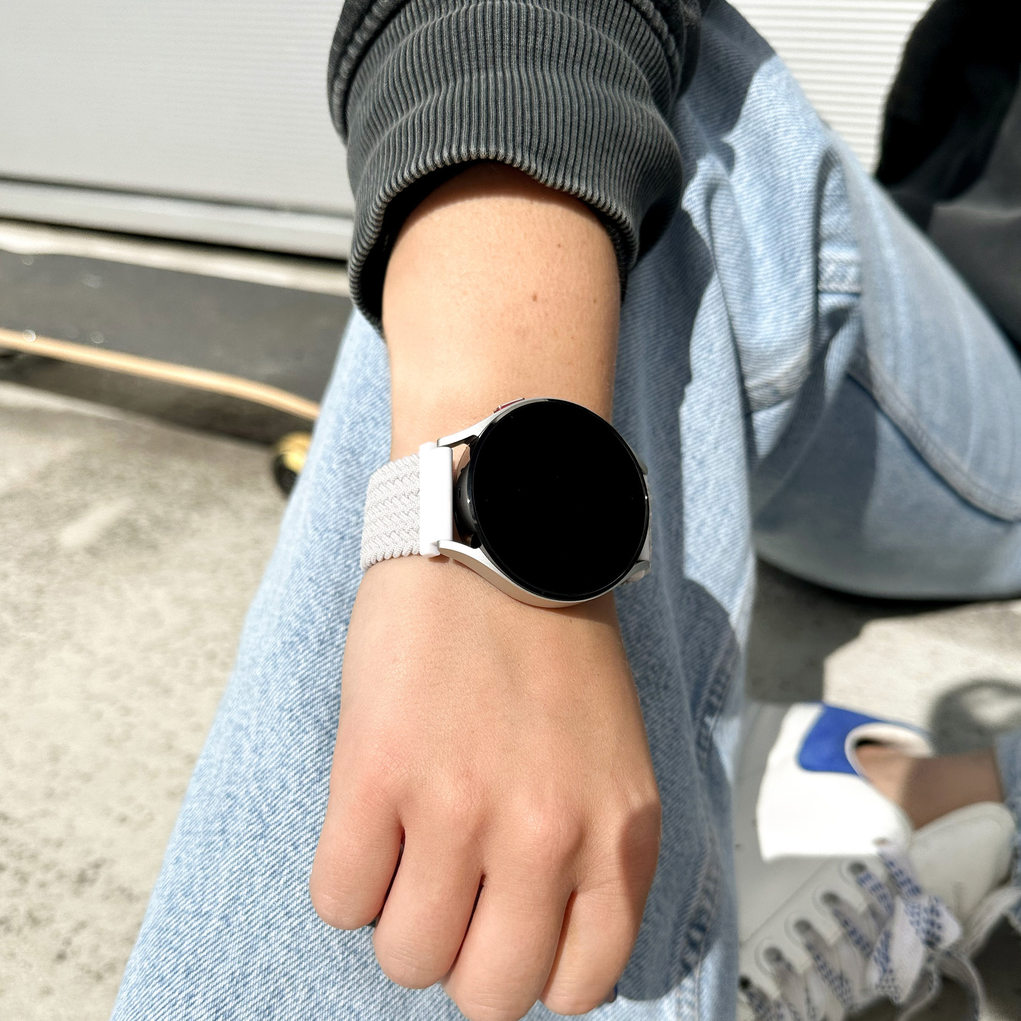 Handgelenk mit silberner Smartwatch, Jeans. Skateboard im Hintergrund.