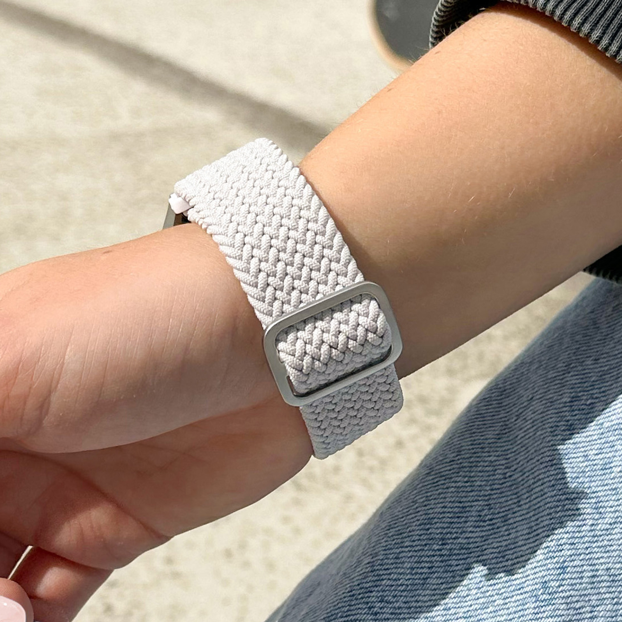 Das Handgelenk einer Person mit einem hellgrauen geflochtenen Uhrenarmband und einer Metallschnalle.