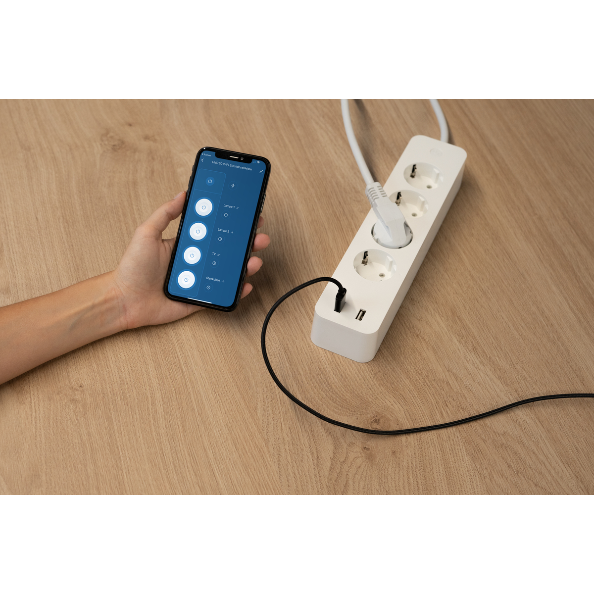 Hand, die ein Telefon hält, das eine intelligente Steckdosenleiste steuert. Auf Holzoberfläche.