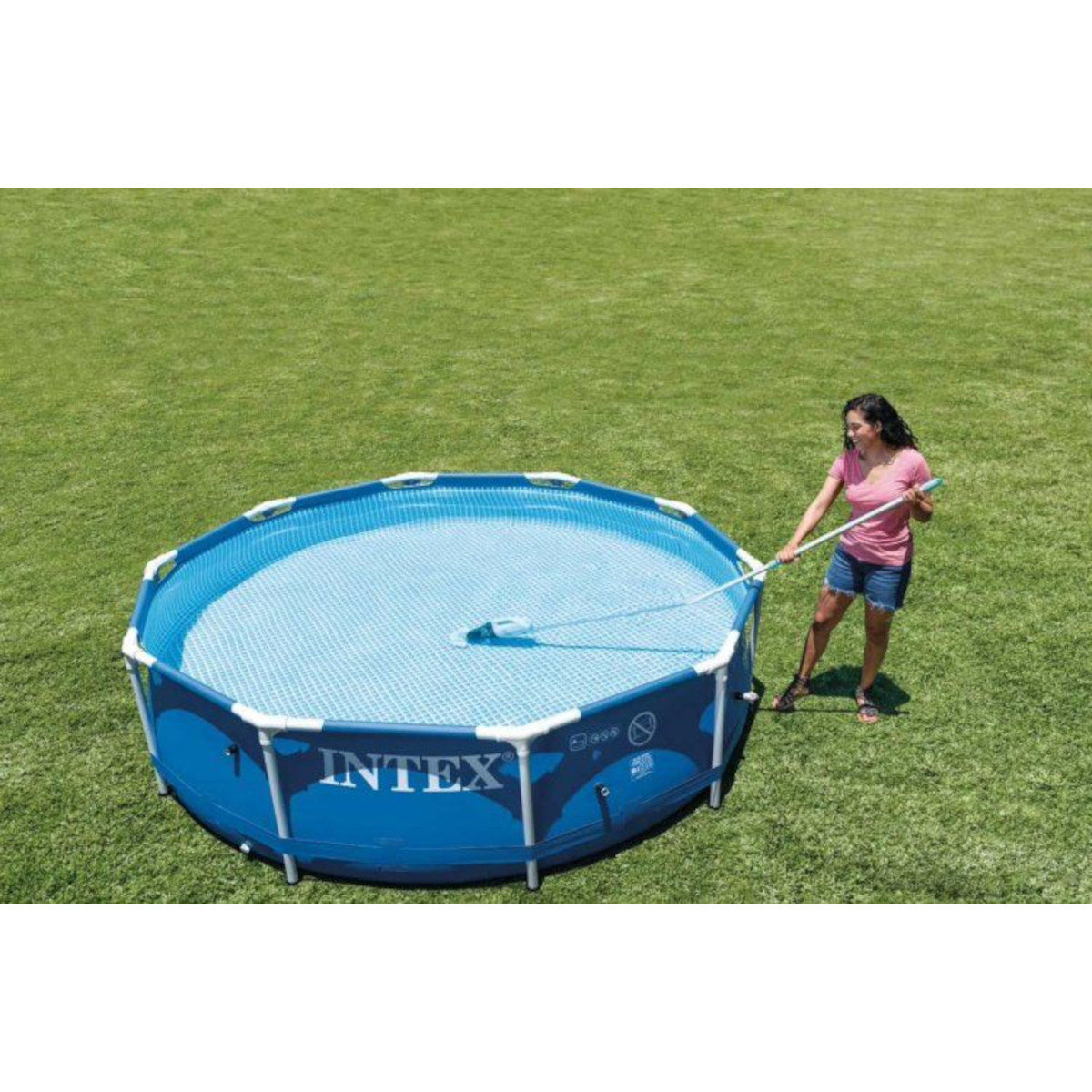 Kobieta czyści niebieski basen naziemny długim kijem, na zielonej trawie, słoneczny dzień.