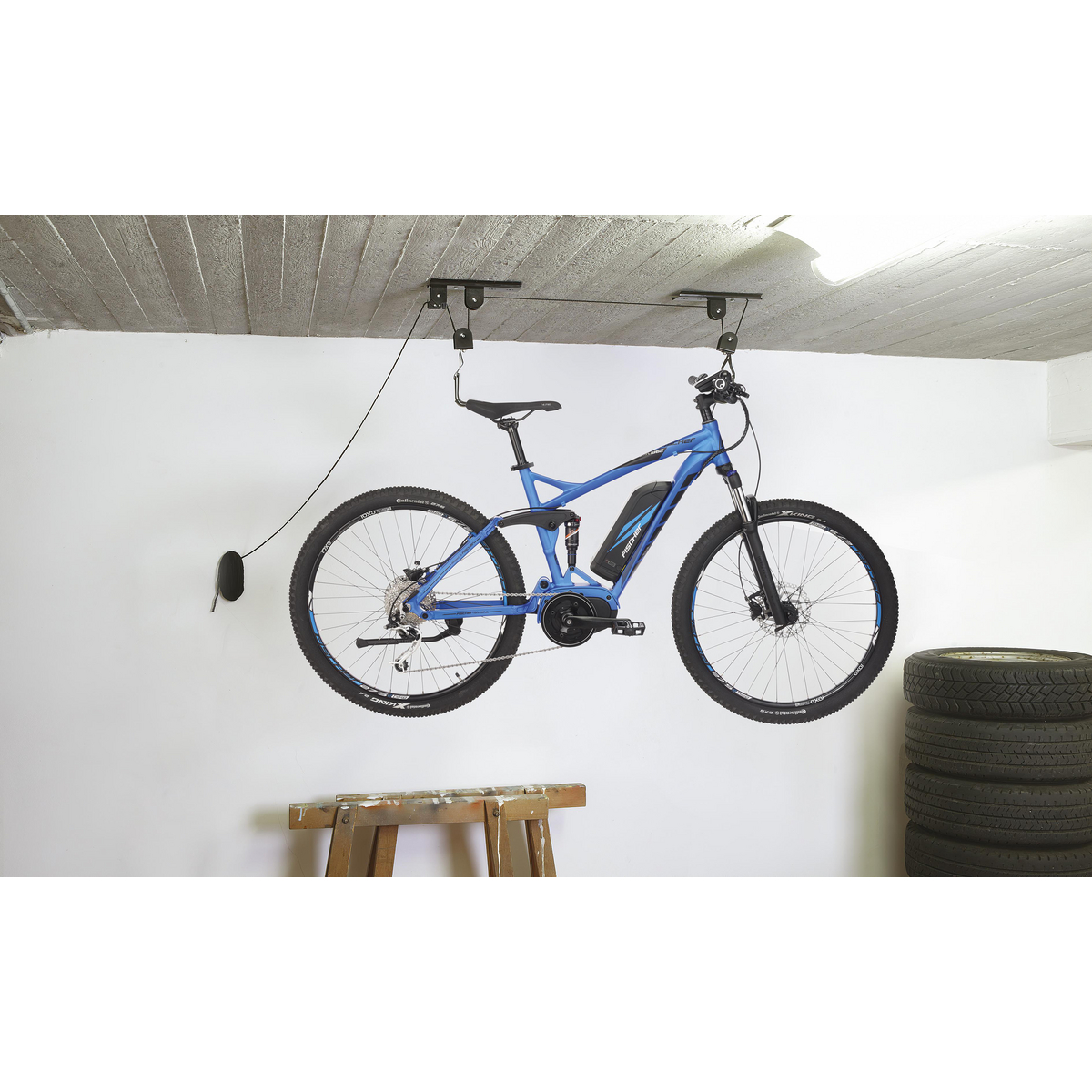 Blaues Fahrrad hängt von der Decke. Weiße Wand, Betondecke, Holzständer, Reifen.