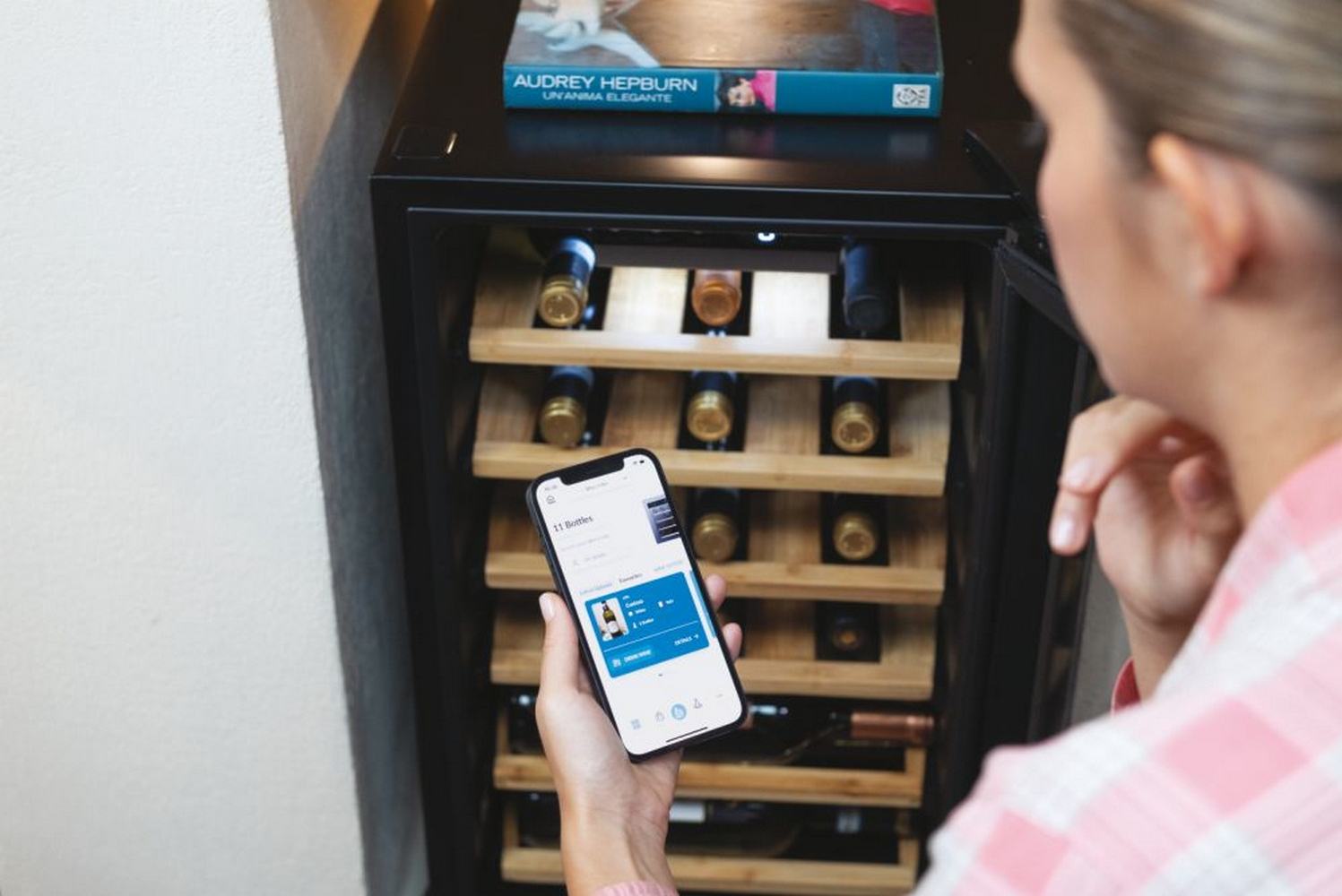 Donna con telefono davanti a una cantinetta aperta. Bottiglie visibili sugli scaffali.