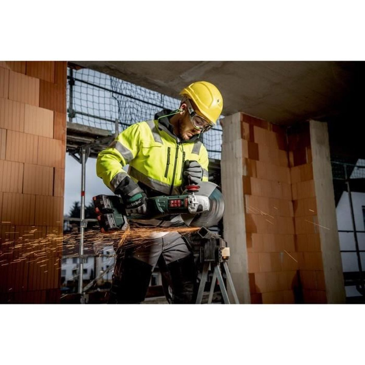 Homme en veste jaune utilisant une meuleuse, des étincelles jaillissent. L'arrière-plan montre la construction.