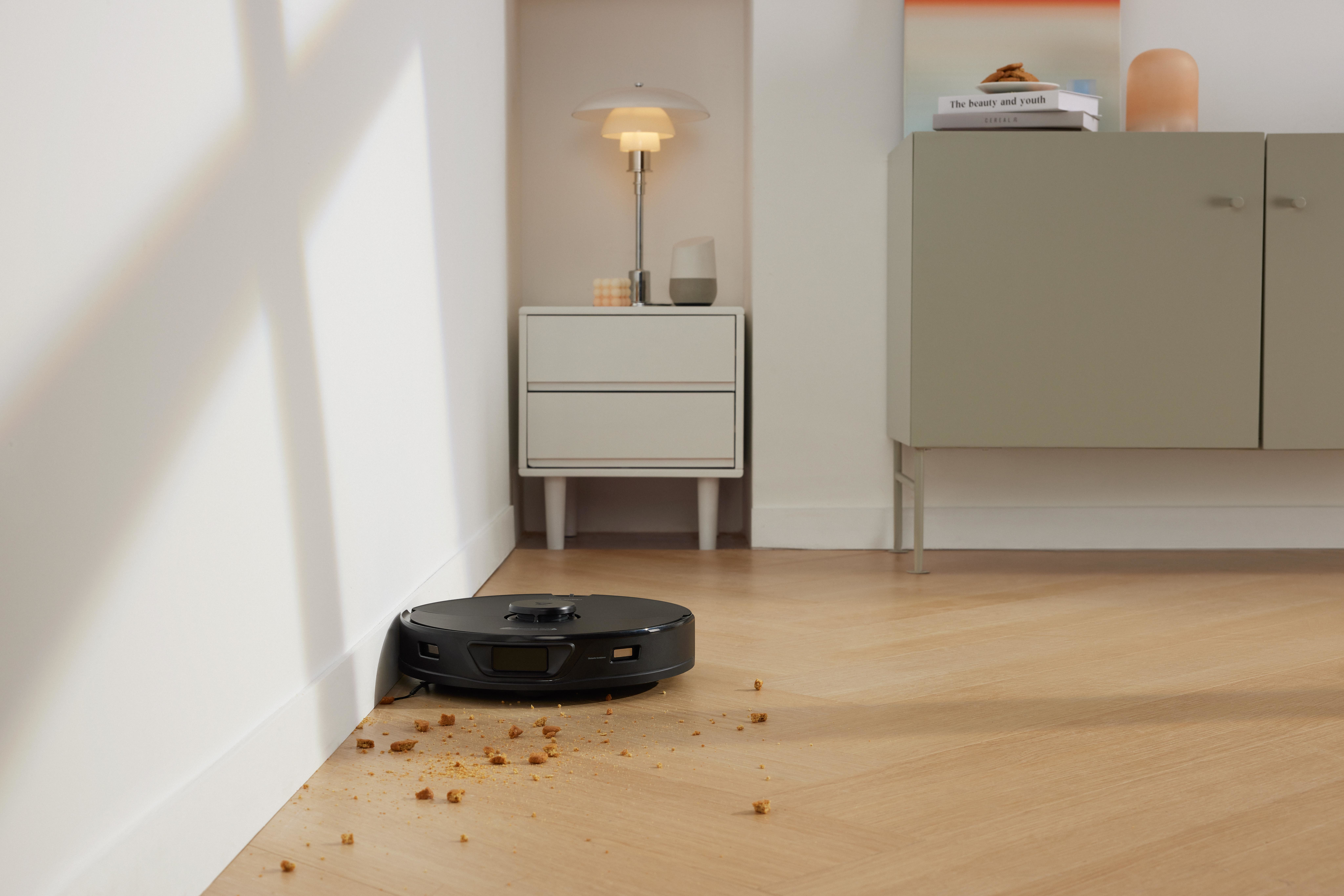 Robot odkurzający czyści drewnianą podłogę. Przed ścianą leżą okruchy.