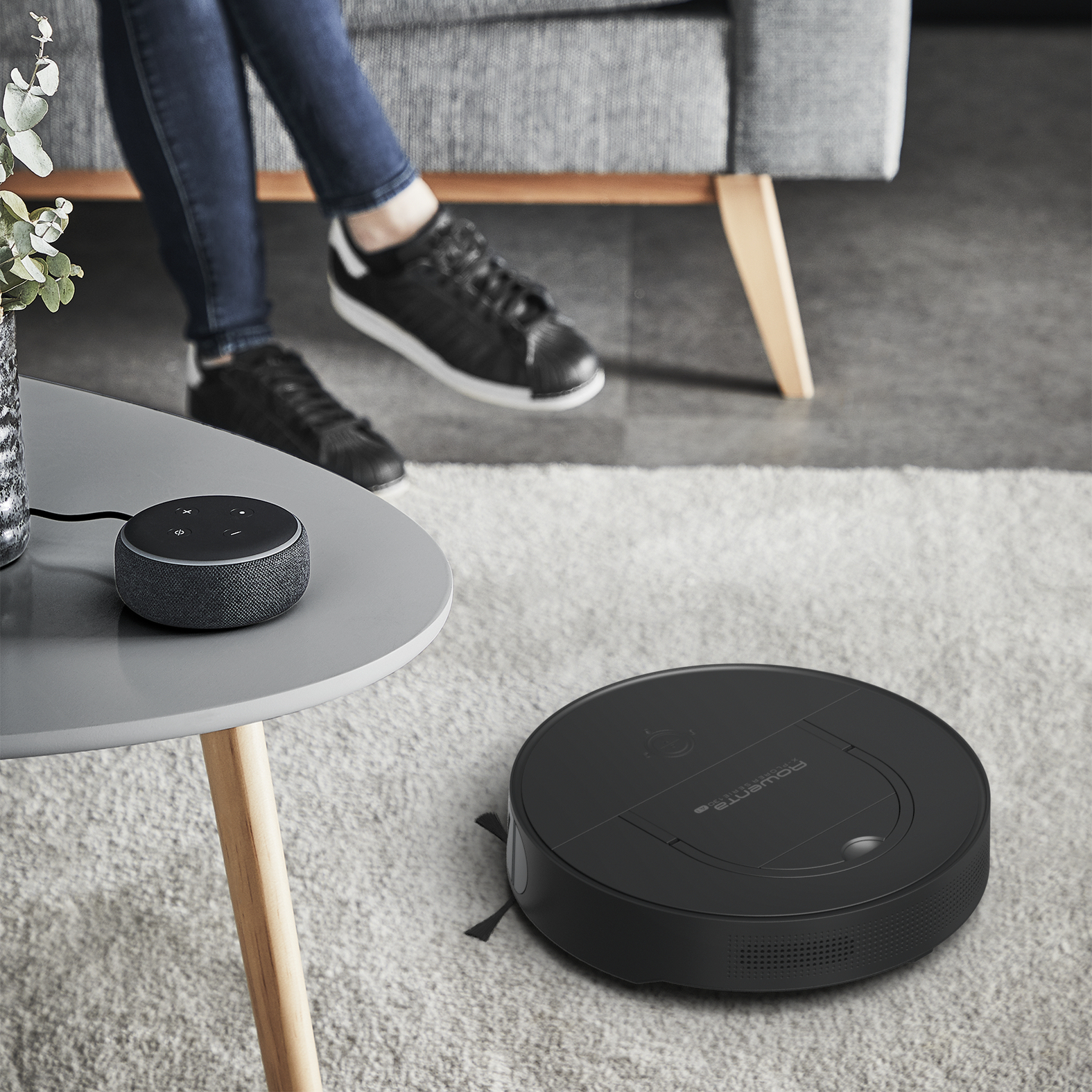 Robot odkurzający na dywanie, z głośnikiem smart i osobą w tle.