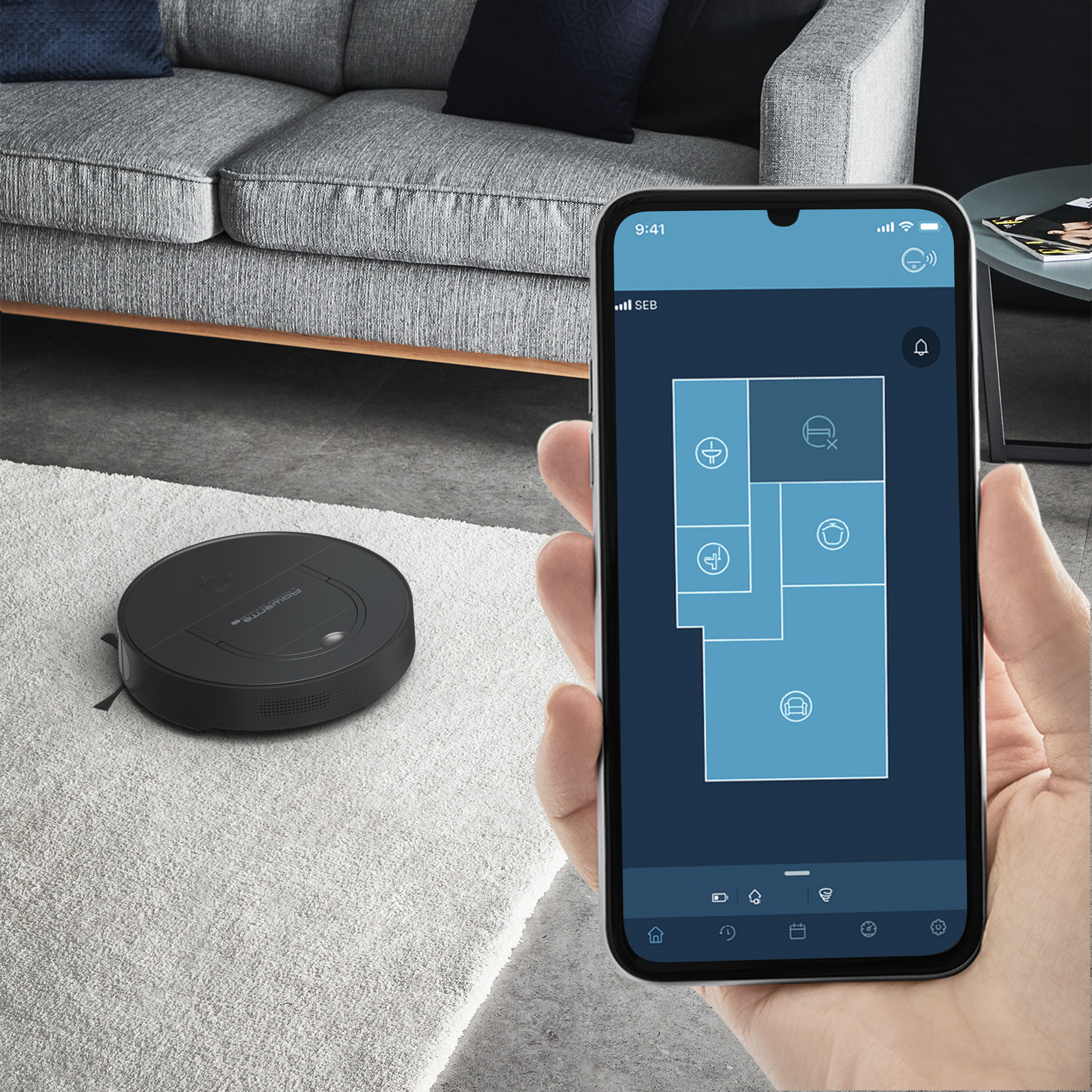 Ręka trzymająca telefon wyświetlający mapę domu. Czarny robot odkurzacz na białym dywanie.