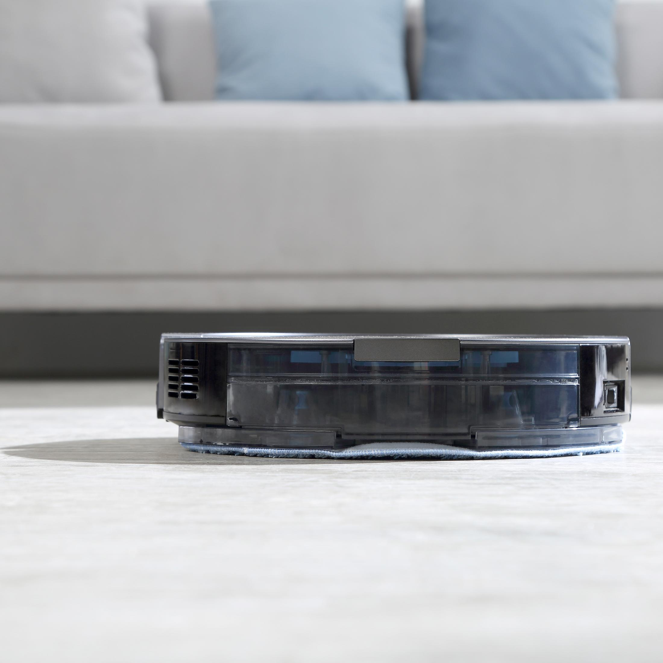 Roboter-Staubsauger auf dem Boden. Ein Sofa und Kissen befinden sich im verschwommenen Hintergrund.