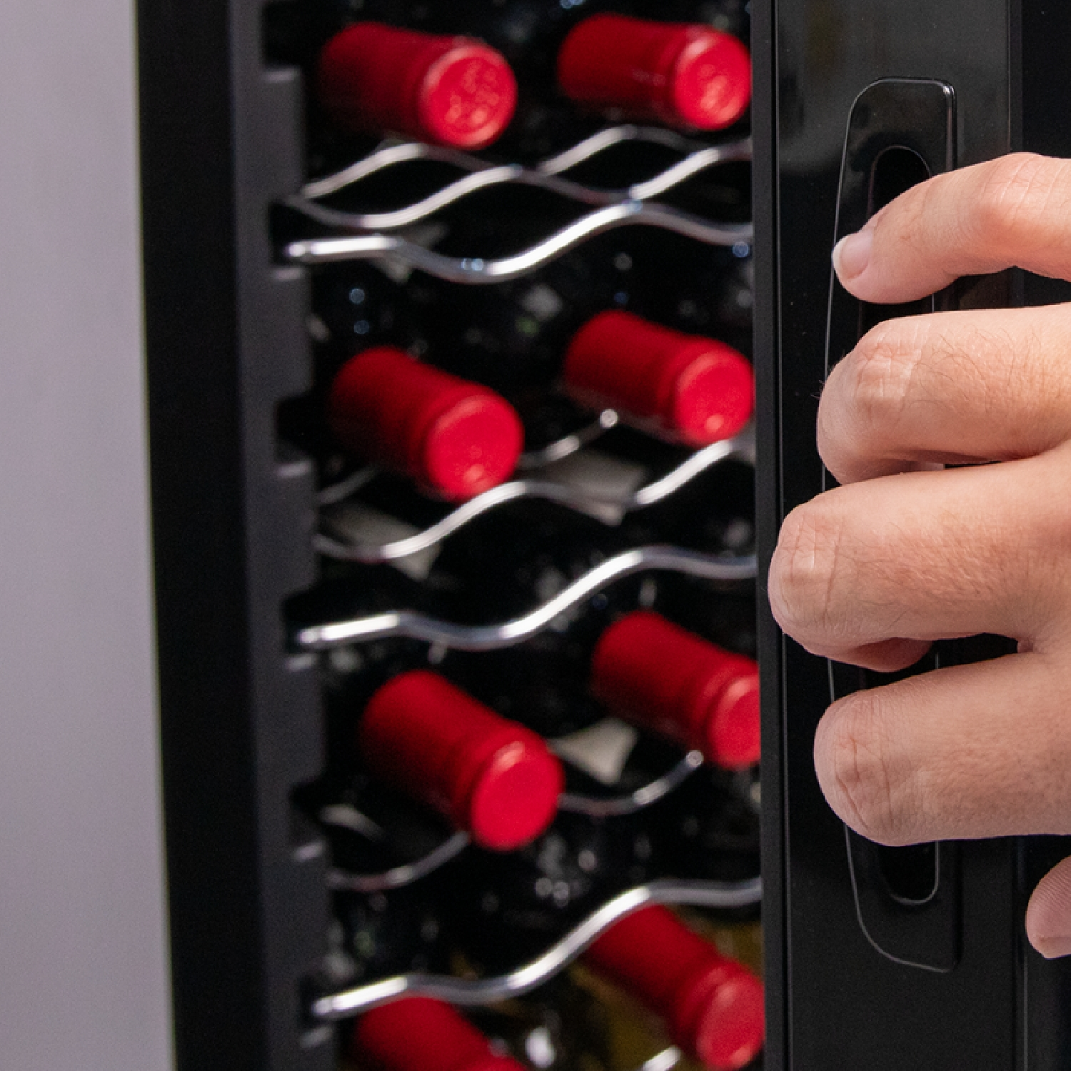 Una mano apre un frigo per vini nero, rivelando bottiglie con tappi rossi.