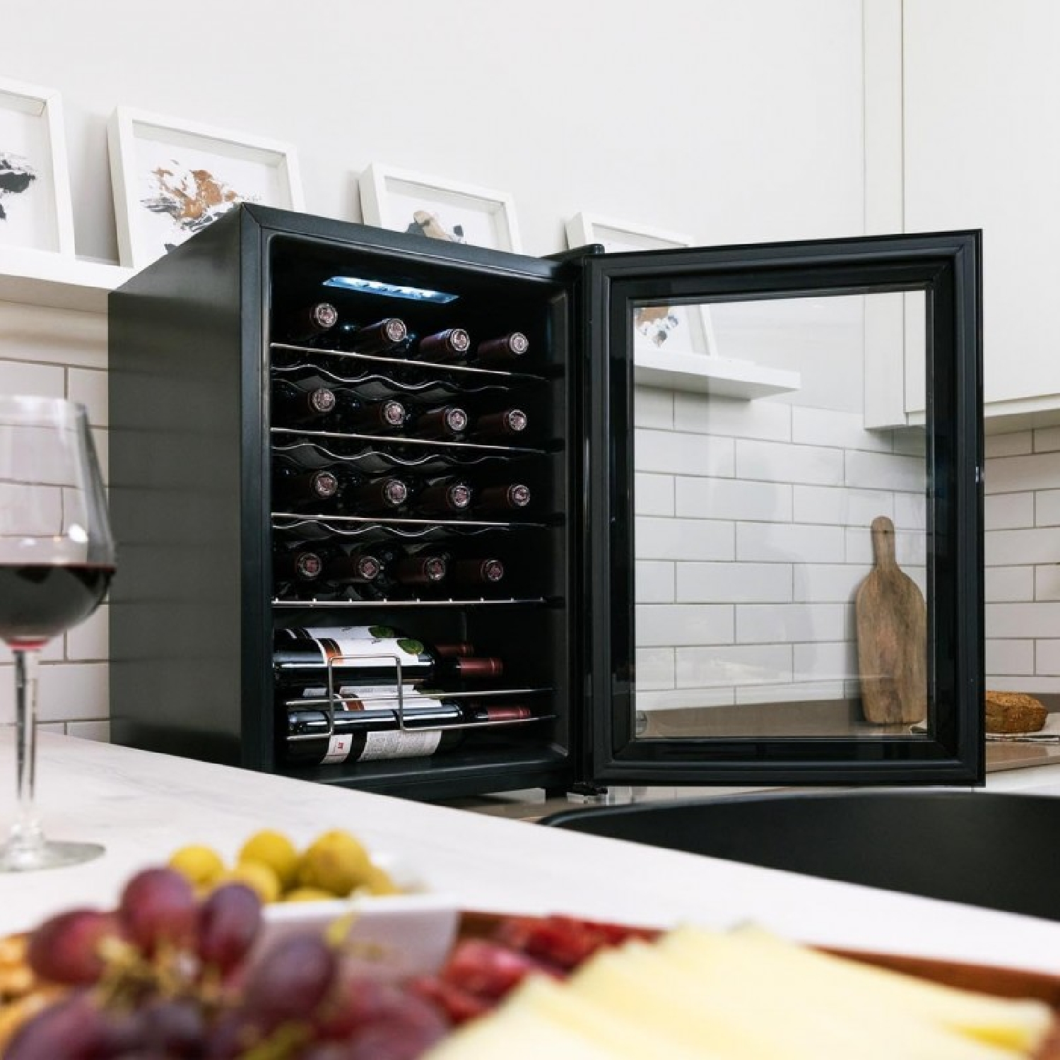 Enfriador de vino con la puerta abierta. Botellas de vino dentro. Copa de vino tinto y uvas en primer plano.