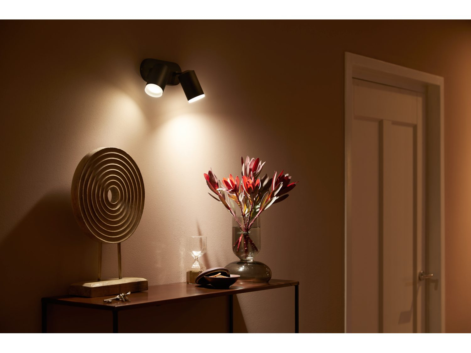 Twee zwarte spotlights op een beige muur, die een tafel met decoratie verlichten. Er is een vaas met bloemen en een witte deur.