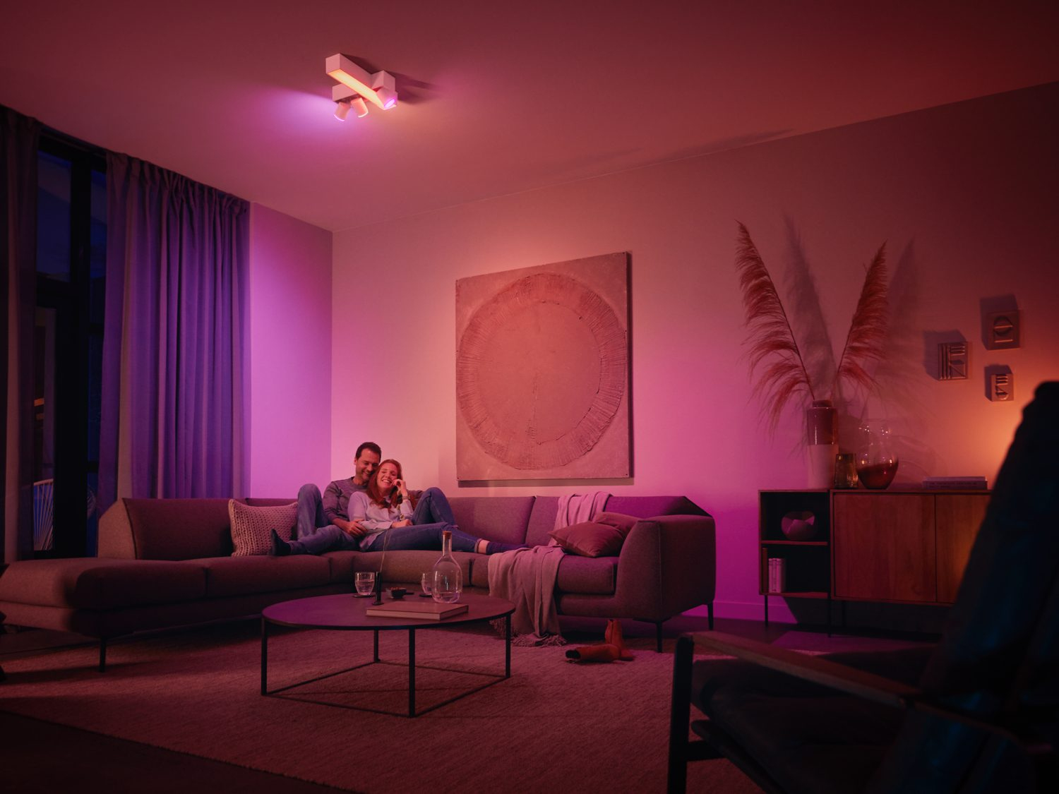 Koppel op een grijze bank in een roze verlichte kamer, met een schilderij en een houten kast.