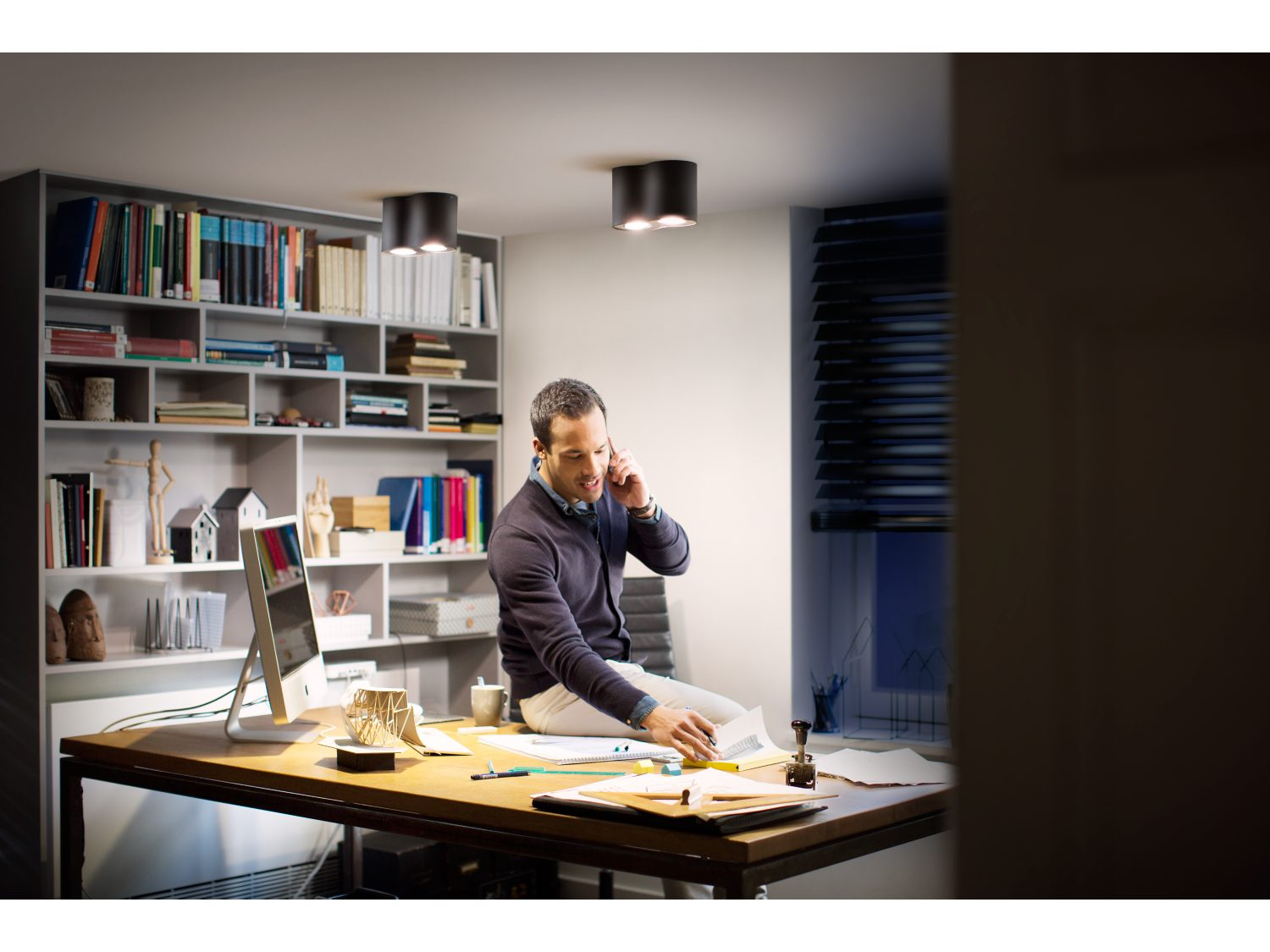 Mężczyzna na biurku z telefonem, laptopem i dokumentami pod oświetleniem sufitowym.