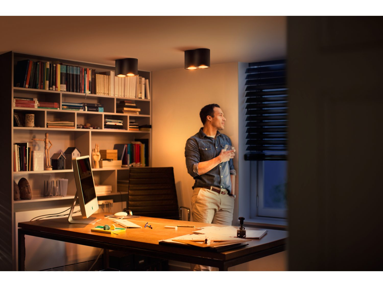 Mężczyzna w biurze patrzy przez okno, biurko z komputerem, regały z książkami w tle.