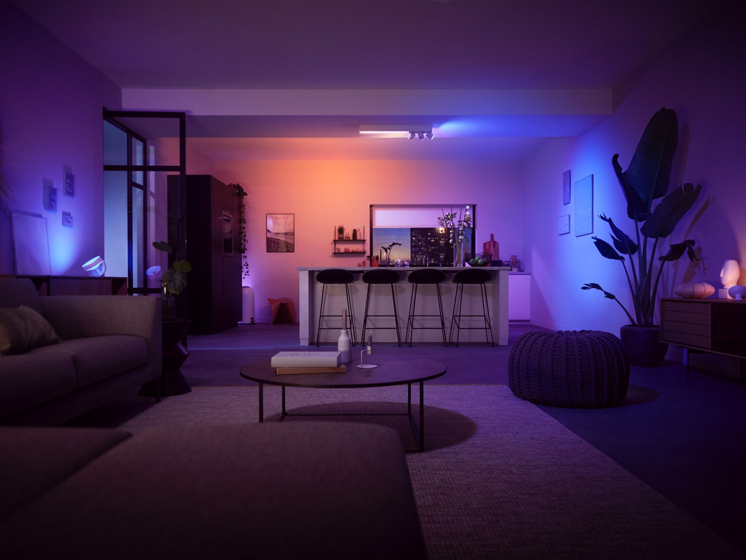 Woonkamer met open keuken. Paarse en oranje sfeerverlichting. Bank, tafel, planten en keukeneiland.