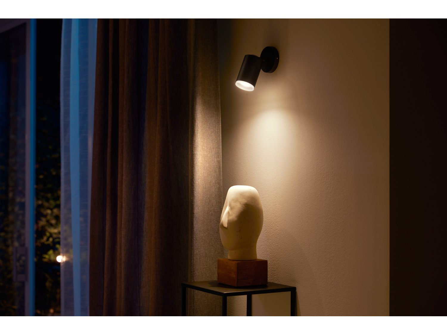Zwarte wandlamp aan een muur. Een sculptuur en een tafel op de voorgrond.