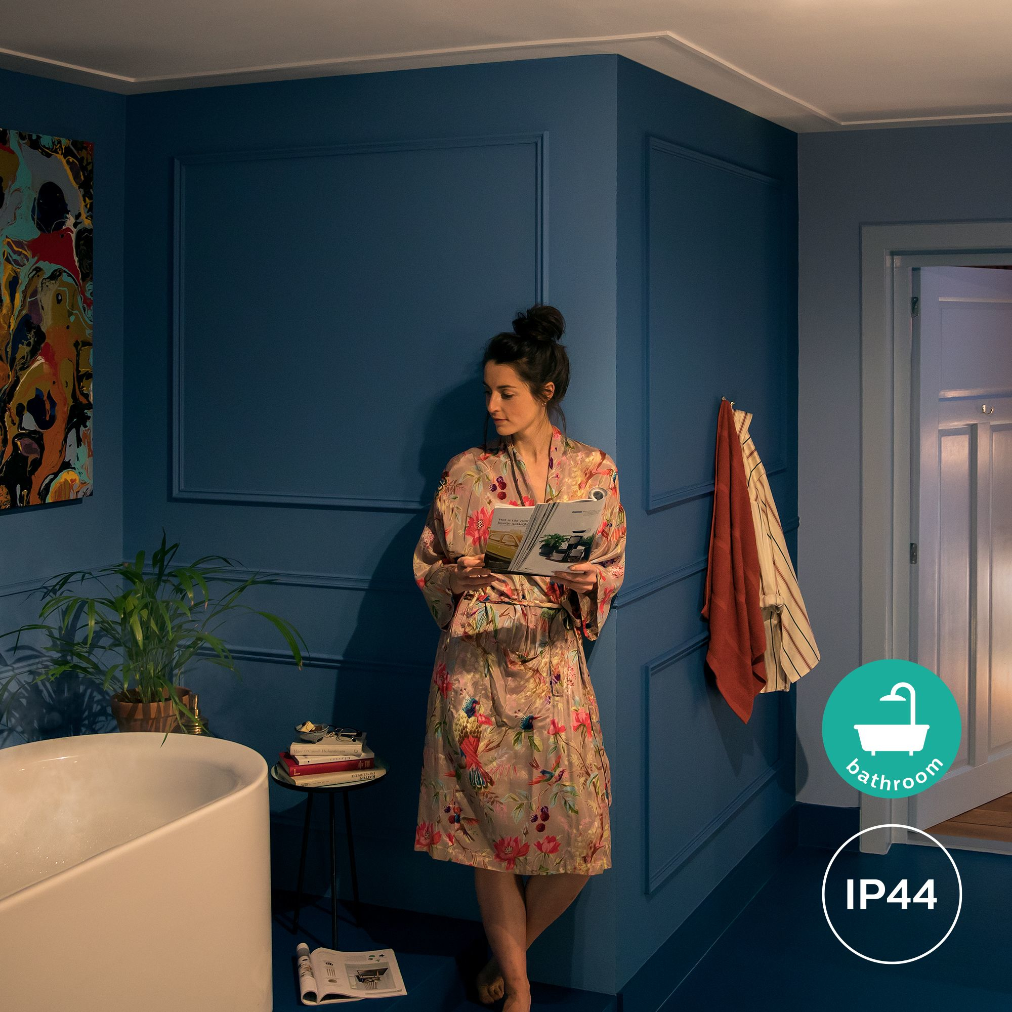 Donna in vestaglia floreale legge in bagno dalle pareti blu. Vasca bianca, dipinto e asciugamano arancione visibili.