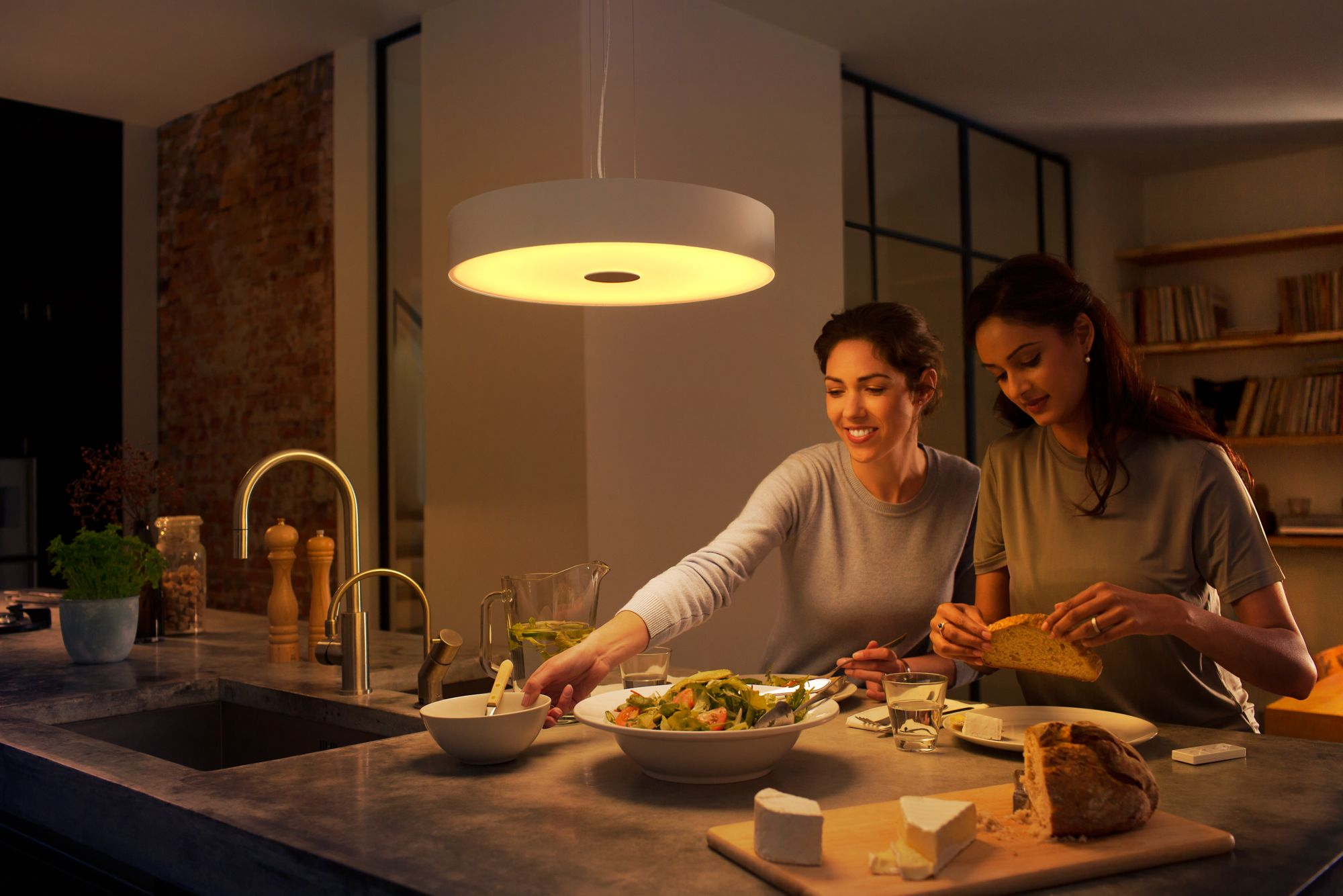Deux femmes dans une cuisine préparant une salade. Une lumière est suspendue. Fromage et pain sont sur le comptoir.