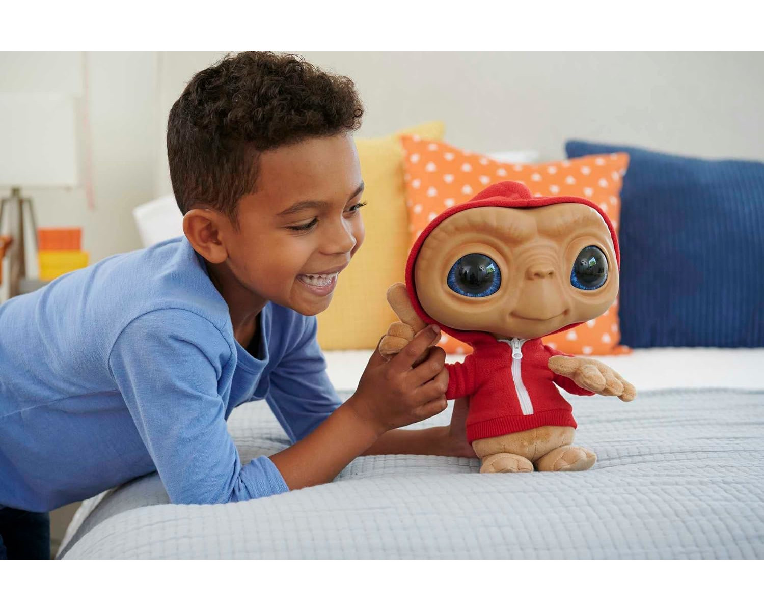 Ein Junge lächelt einen E.T.-Plüschtier an, das einen roten Hoodie trägt. Sie sind auf einem Bett mit bunten Kissen.