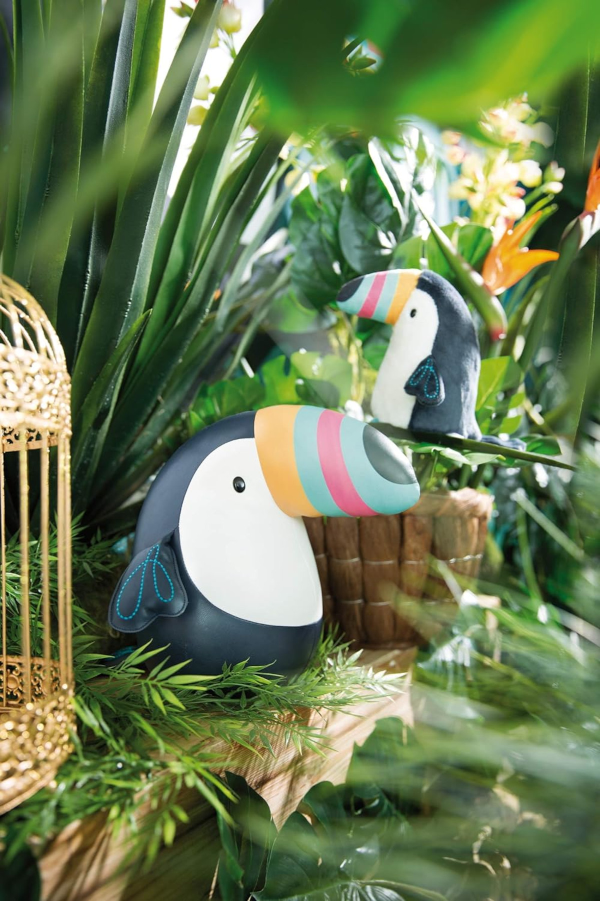 Zwei Tukan-Stofftiere zwischen Blättern und einem goldenen Vogelkäfig. Leuchtende Farben.