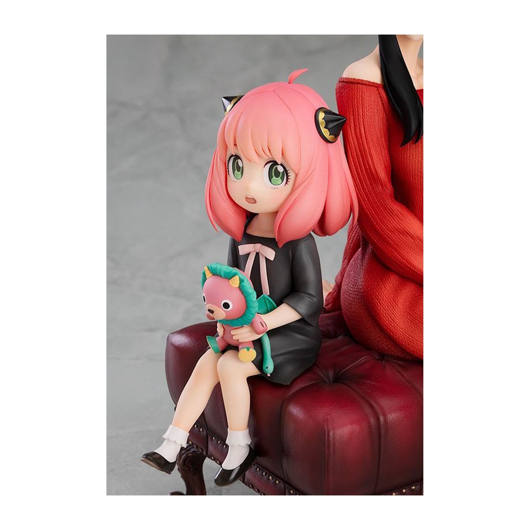 Anime-Mädchenfigur, die ein Spielzeug hält. Trägt ein schwarzes Kleid. Im Hintergrund eine Person in rotem Pullover.