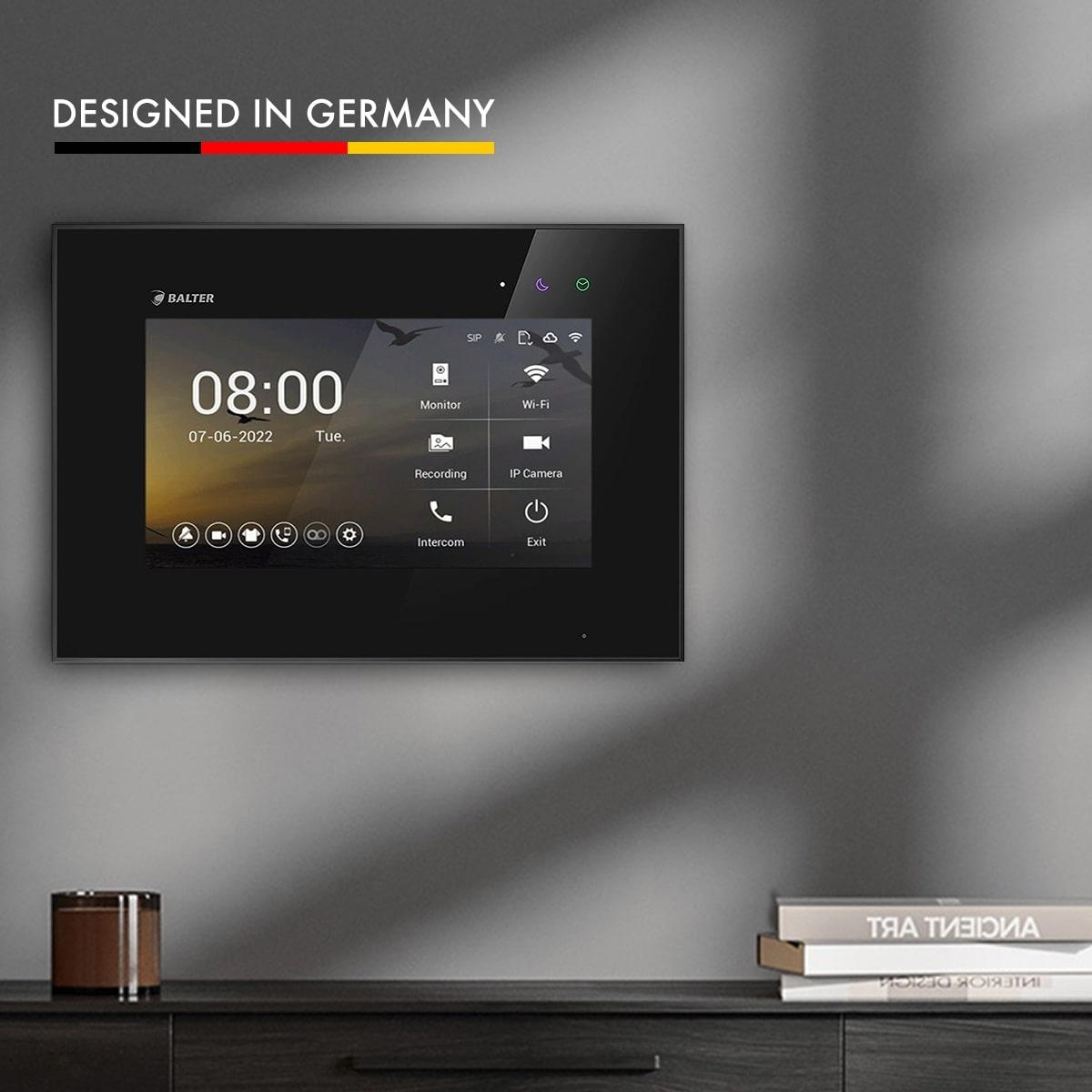 Schwarzer Intercom-Monitor an der Wand, mit Uhr und Symbolen, Text 'Designed in Germany'.