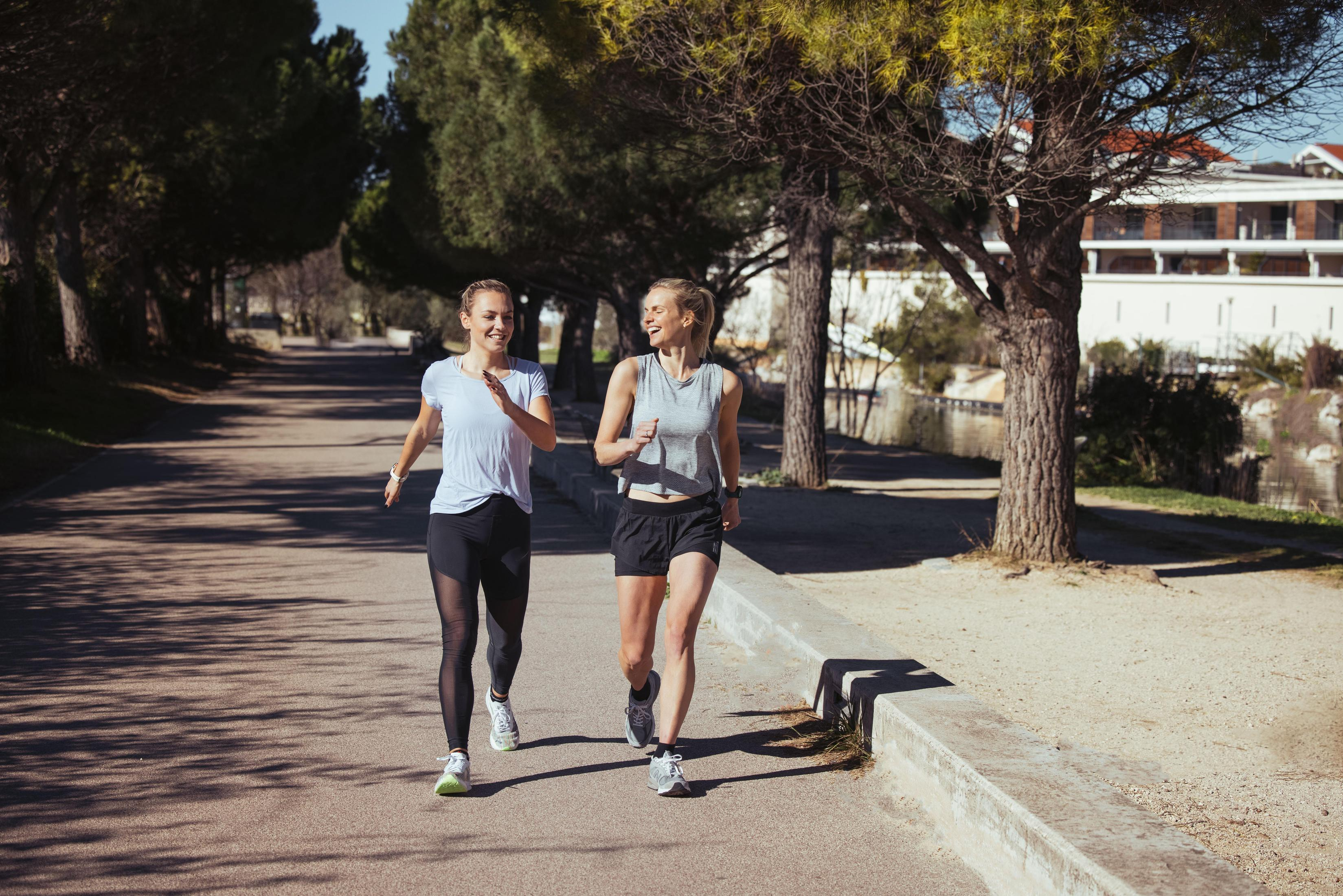 Dwie kobiety biegają ścieżką w parku, drzewa w tle, słoneczny dzień.