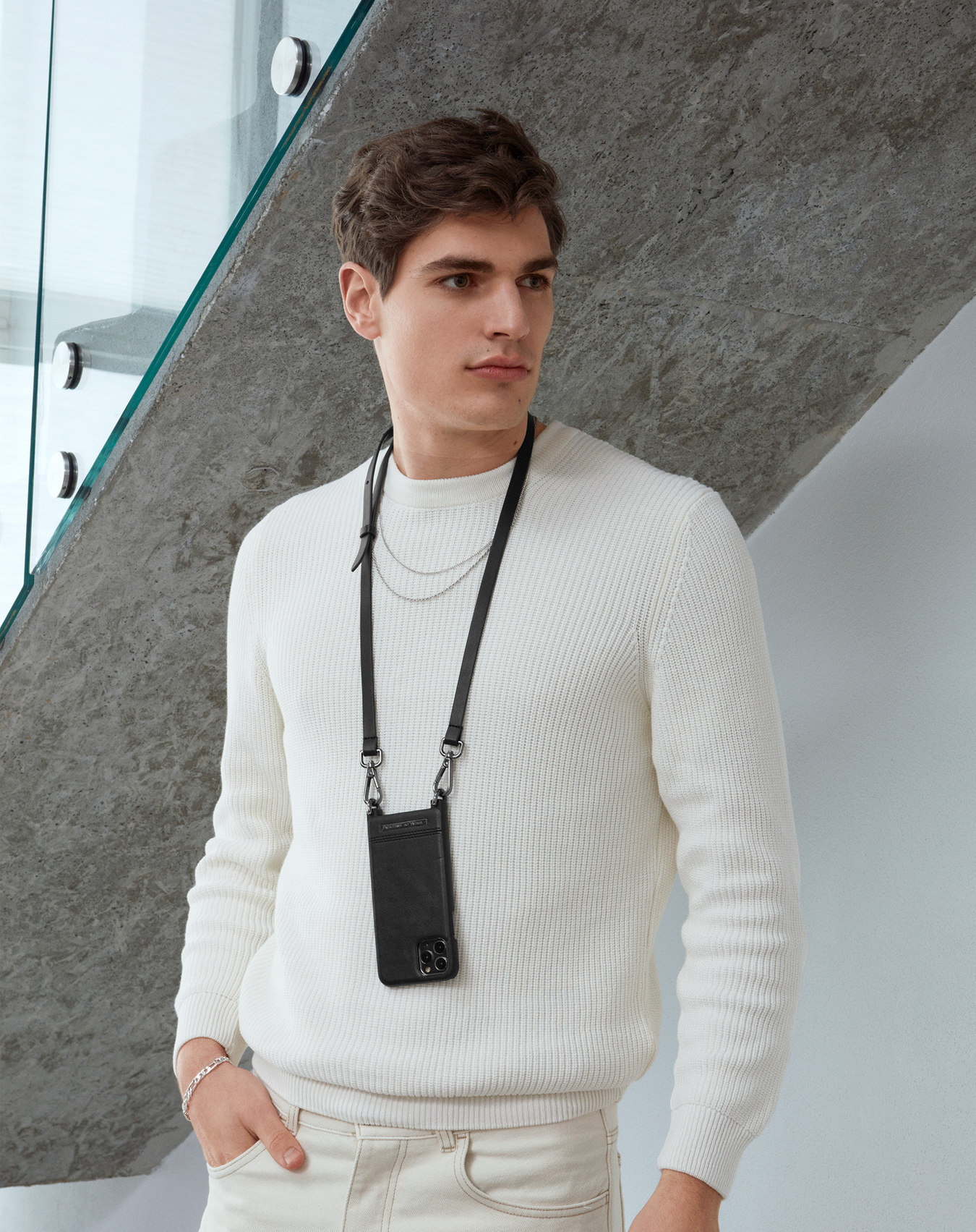 Ein Mann mit cremefarbenem Pullover und schwarzer Handyhülle an einem Riemen. Der Hintergrund ist eine graue Wand.