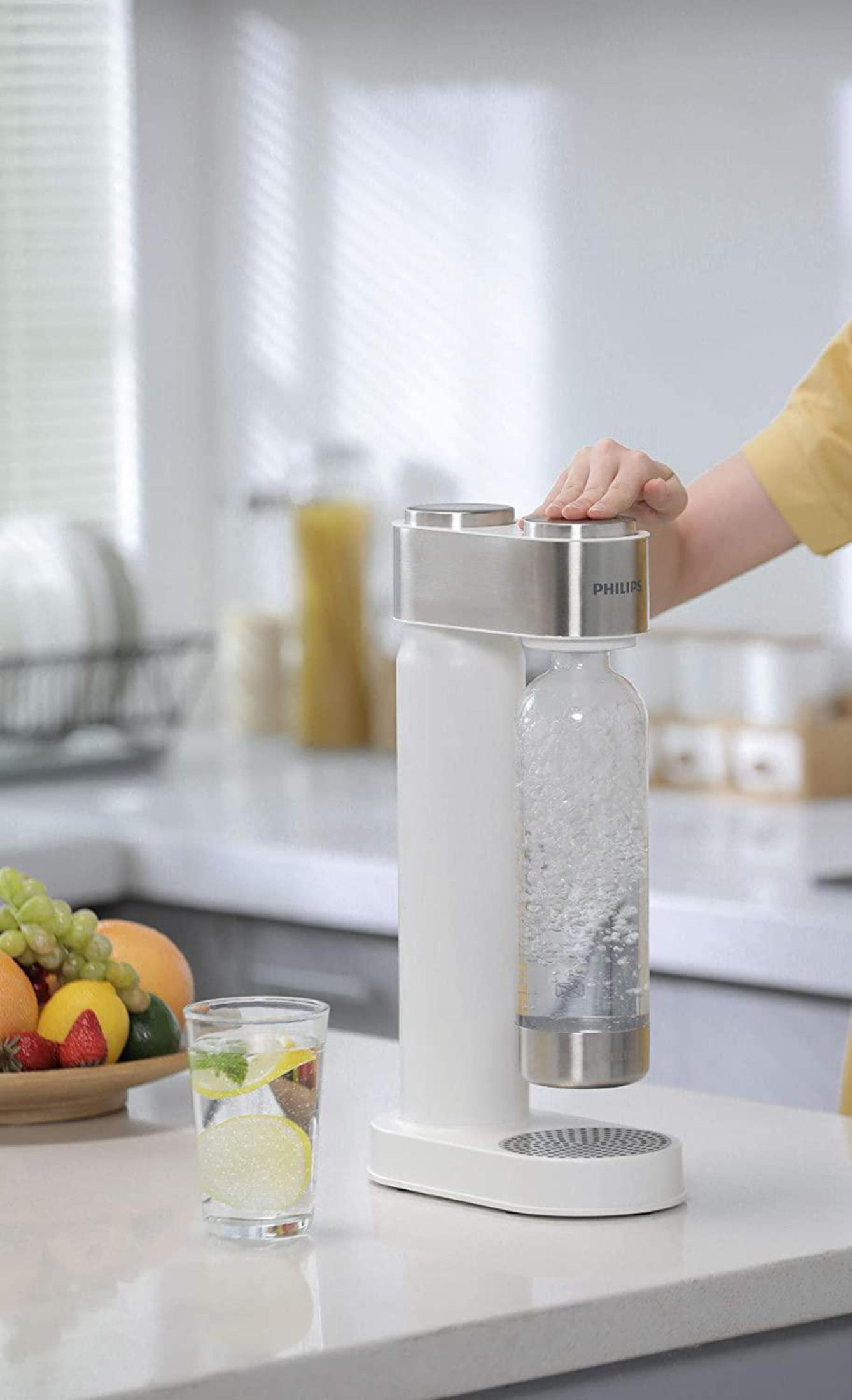 Weißer und silberner Philips Soda Maker, Glas Wasser mit Zitronenscheiben und eine Obstschale.