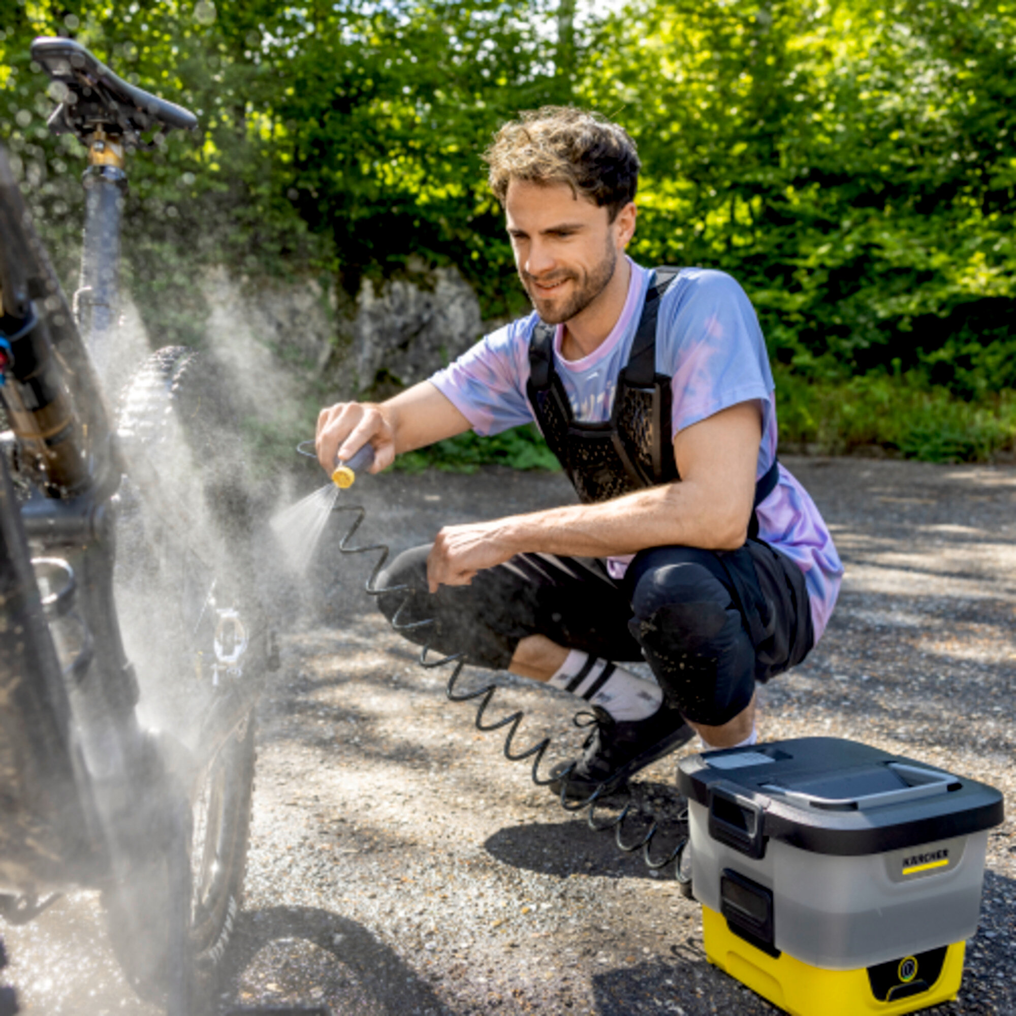 Mann hockt, wäscht Fahrrad mit Schlauch. Wasser spritzt aus dem Schlauch. Gelb-graue Maschine daneben.