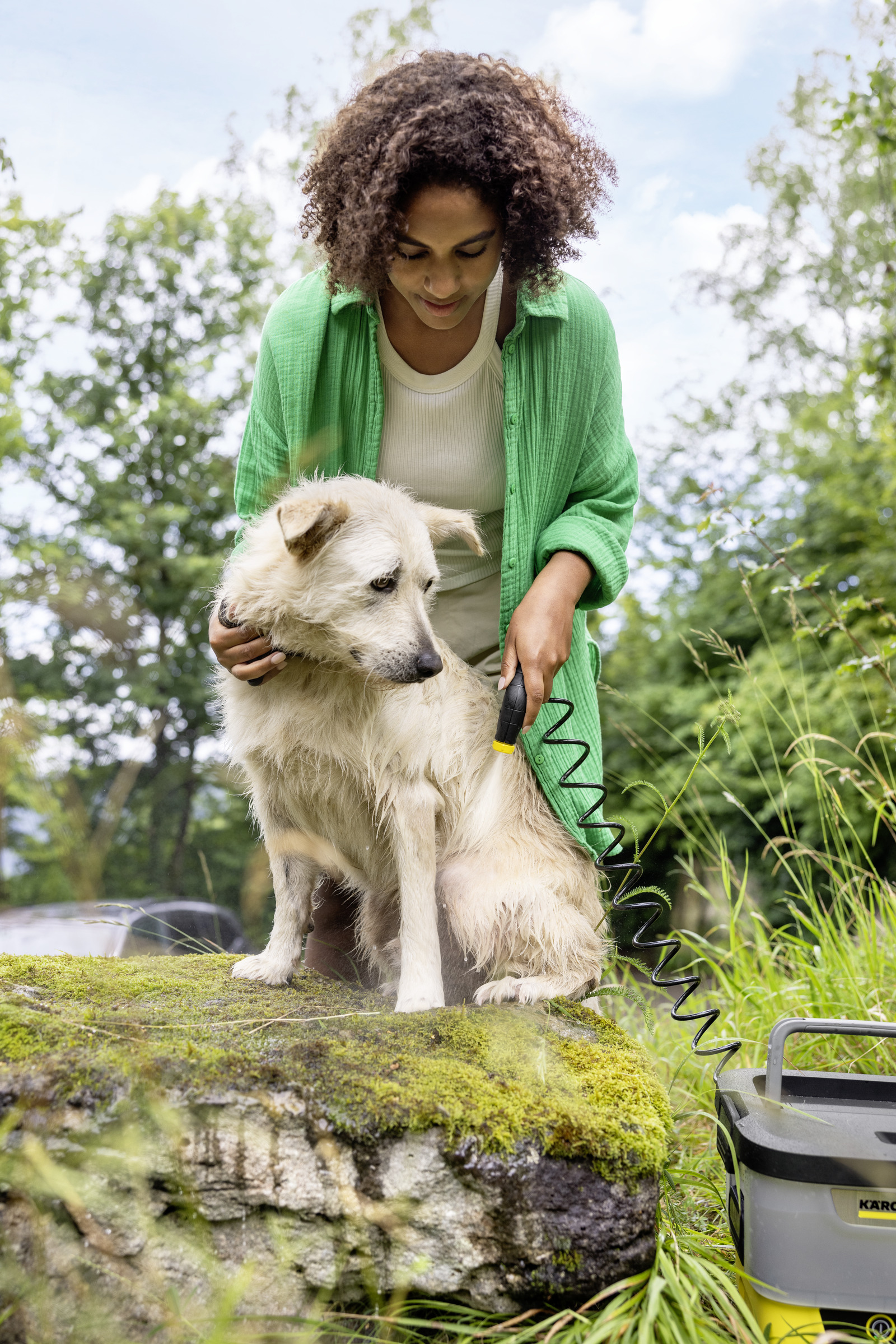 Eine Frau wäscht einen nassen Hund auf einem moosigen Felsen mit einem Schlauch. Grüne Bäume und Himmel im Hintergrund.