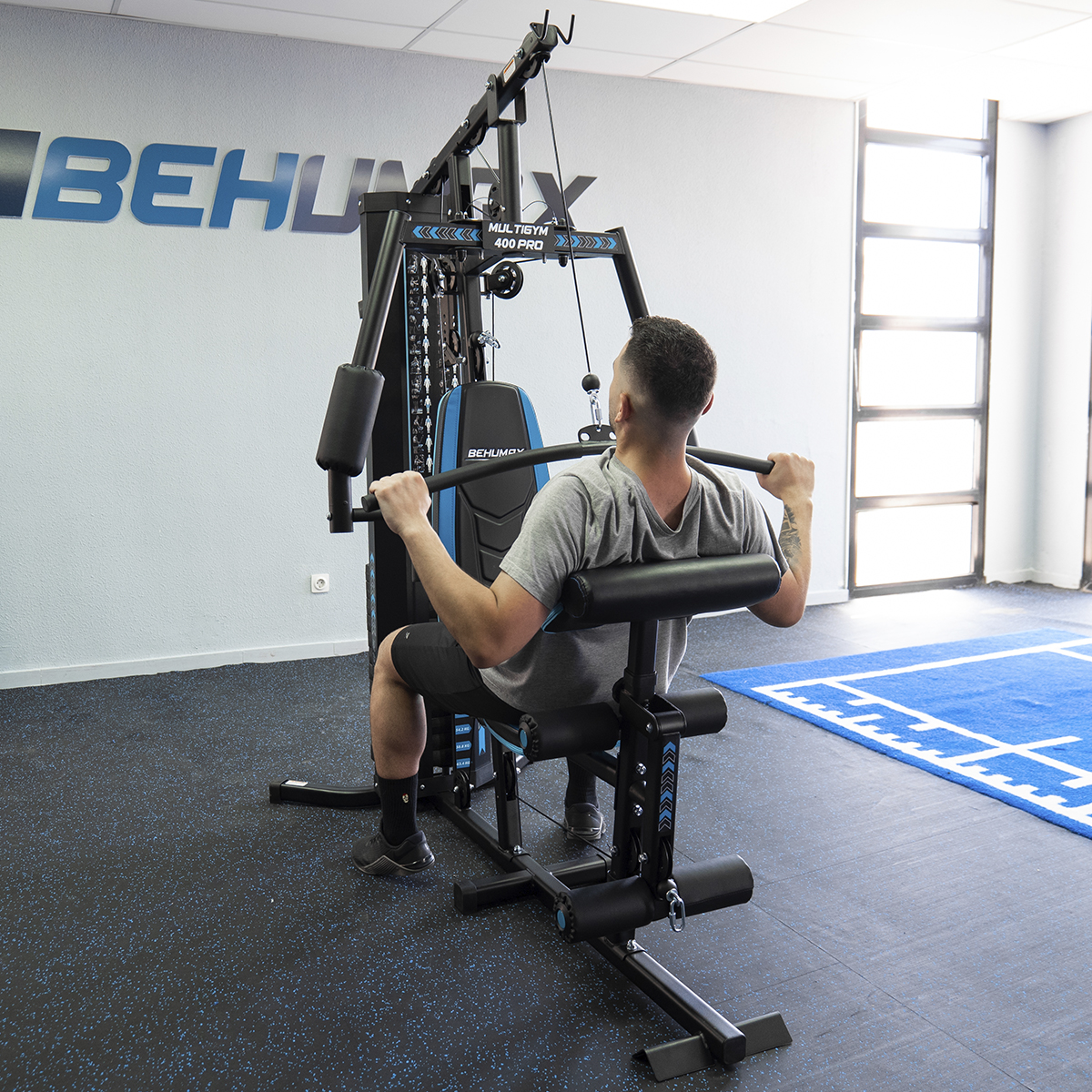 Hombre usando máquina de pesas en gimnasio, con alfombra azul y marca Behumax en la pared.