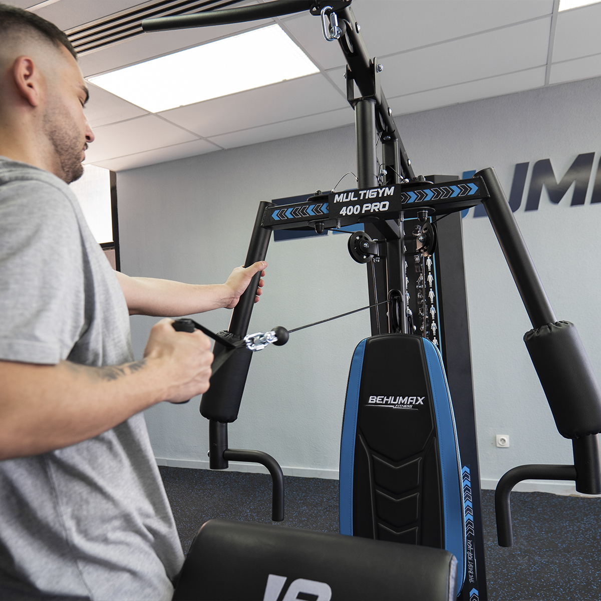 Hombre haciendo ejercicio en un multi-gimnasio. Tira de un cable. El gimnasio es negro y azul con "BEHUMAX" escrito.