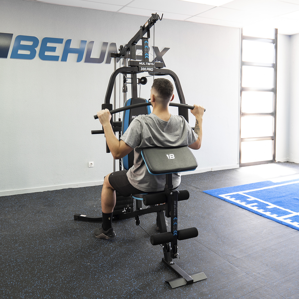 Hombre en gimnasio ejercitándose en una máquina de ejercicios negra y azul. Pared blanca y azul.