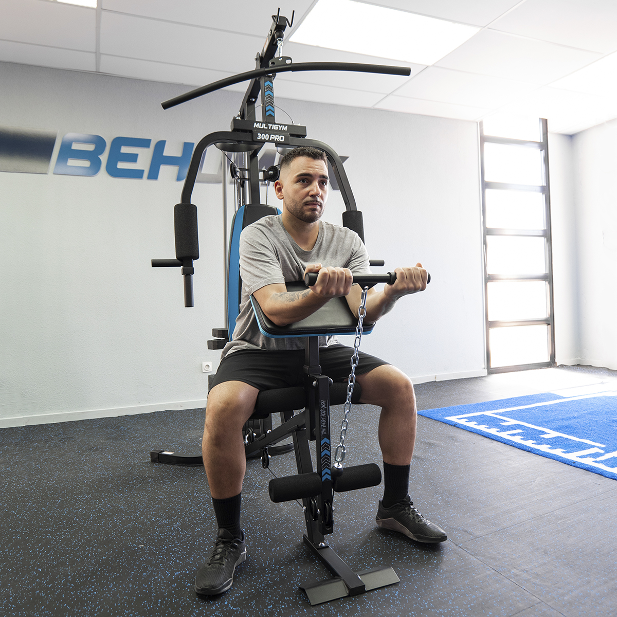 Hombre usando una máquina de gimnasio múltiple. El gimnasio está en una habitación con equipos de ejercicio y una estera azul.