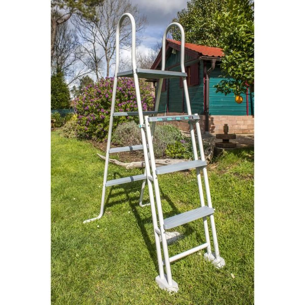 Weiße Metallleiter in einem grünen Garten, ein kleines Haus mit rotem Dach im Hintergrund.