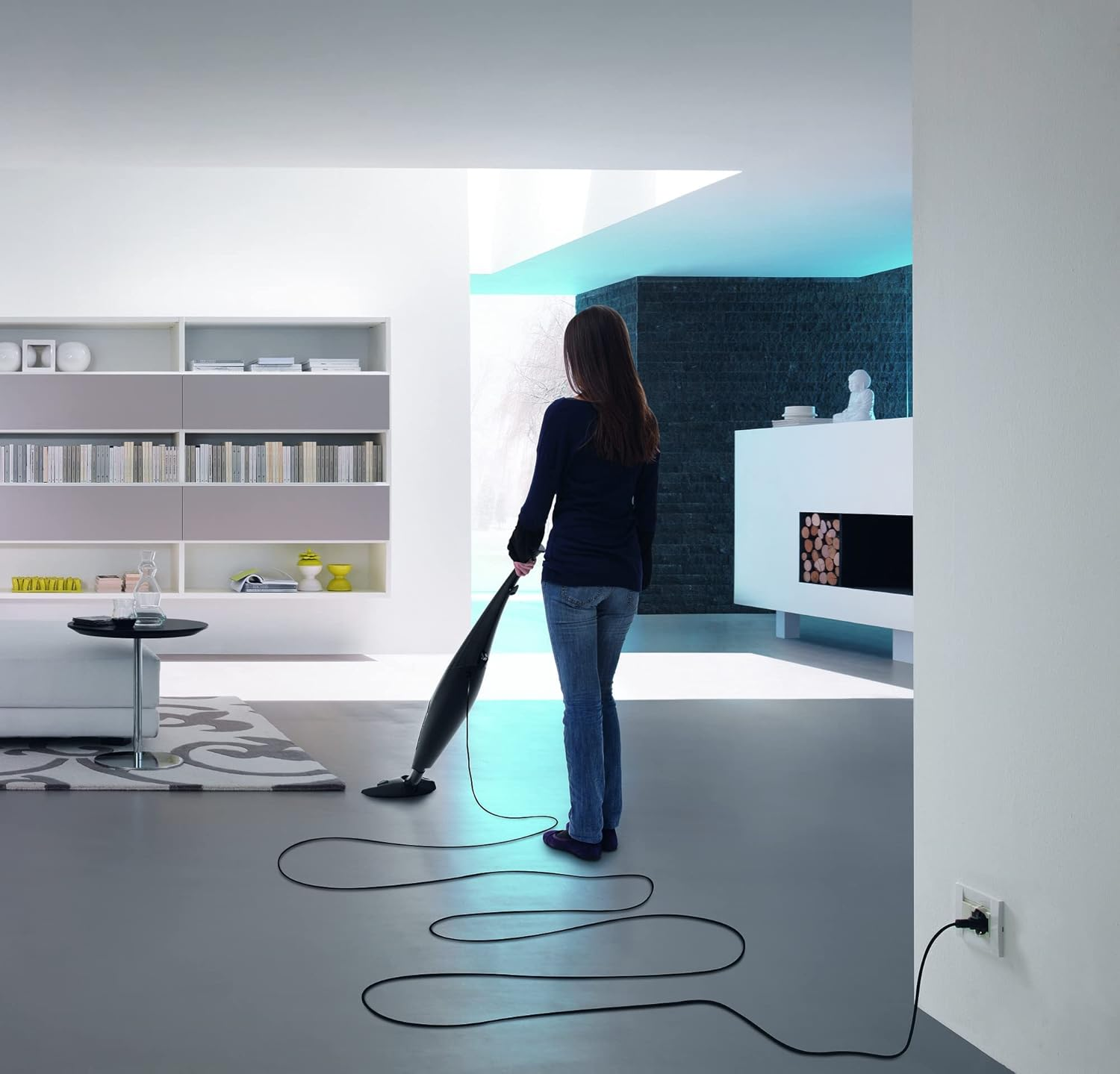 Kobieta odkurza szarą podłogę w nowoczesnym, białym pokoju. Kabel jest rozłożony po podłodze.