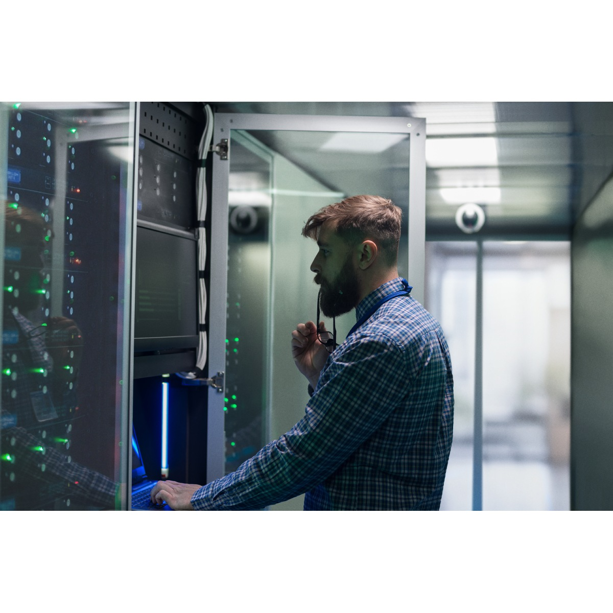 Mężczyzna w koszuli w kratę pracuje na laptopie w serwerowni, trzymając okulary i patrząc na ekran.