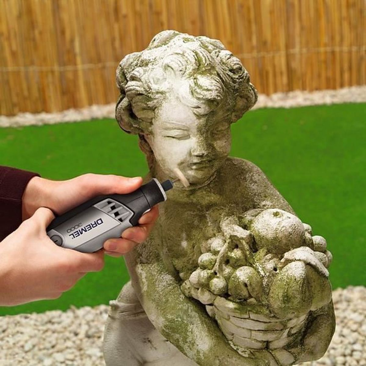 Hände benutzen ein Dremel-Werkzeug an einer Statue, mit grünem Hintergrund aus Gras und Hecke.