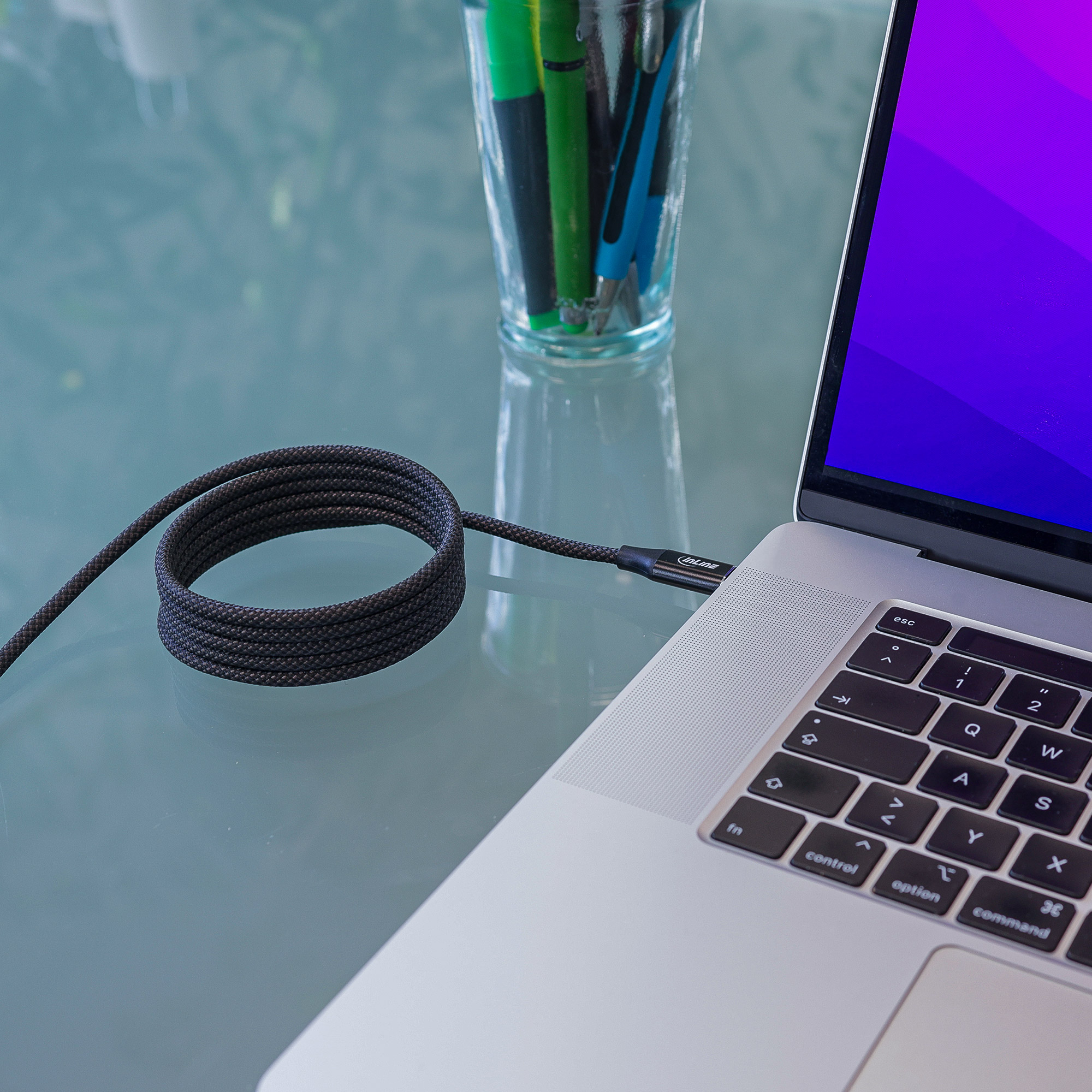 Câble USB-C tressé noir connecté à un ordinateur portable. Le câble forme une boucle, à côté de stylos.