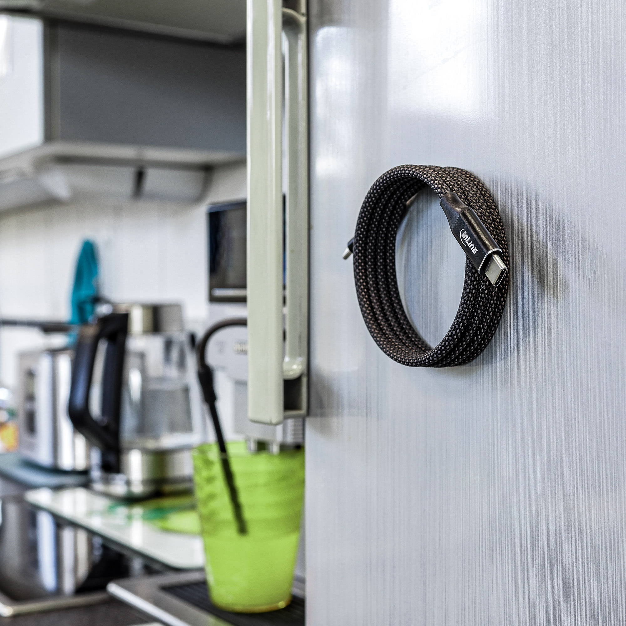 Câble USB-C noir enroulé sur un réfrigérateur argenté. Tasse verte et cuisine en arrière-plan.