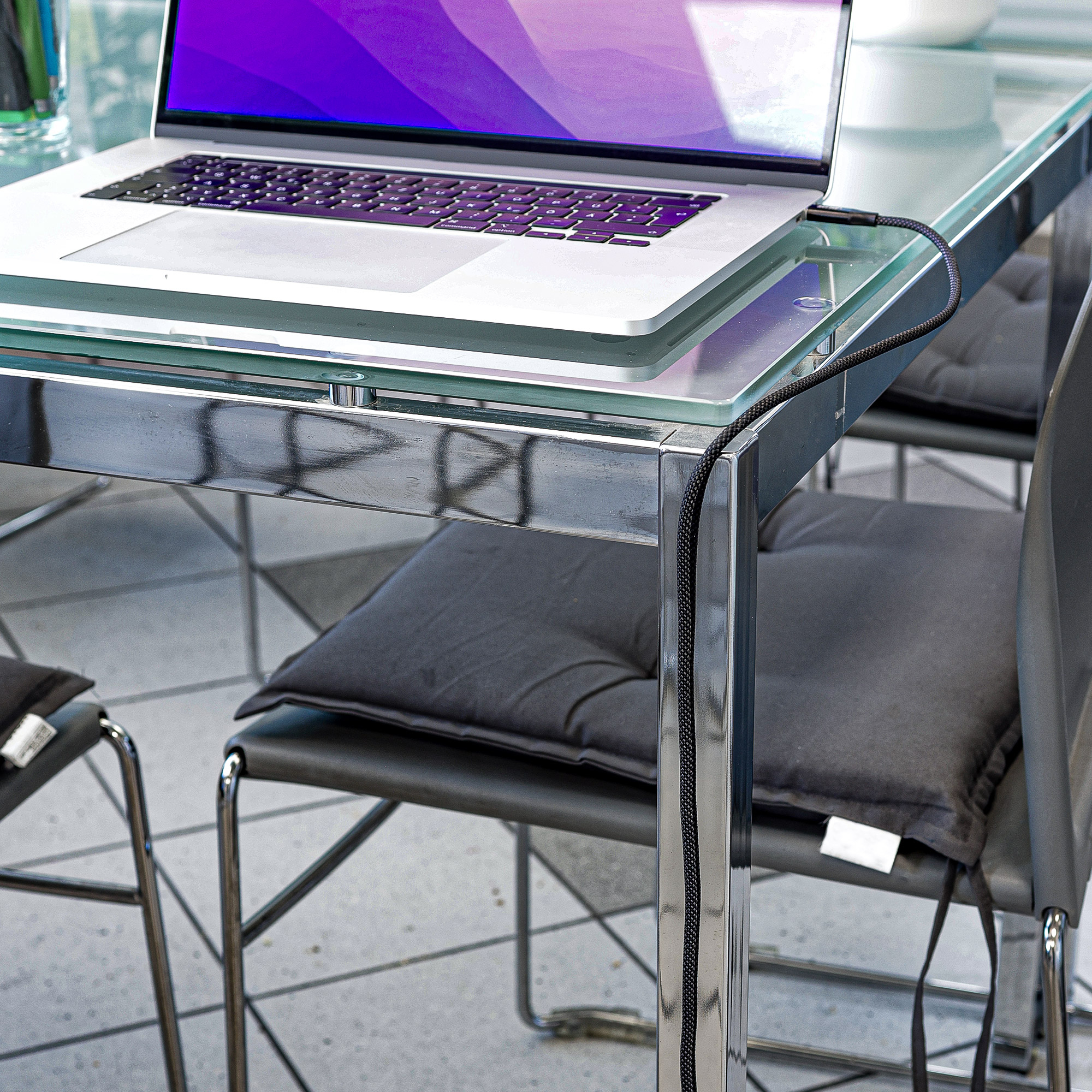 Un ordinateur portable sur une table en verre, connecté par un câble noir à une chaise noire. Écran violet.