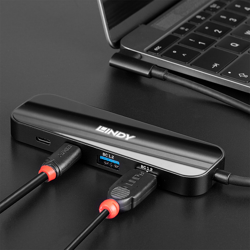 Hub nero con porte, cavi USB collegati, su una scrivania con un laptop sullo sfondo.