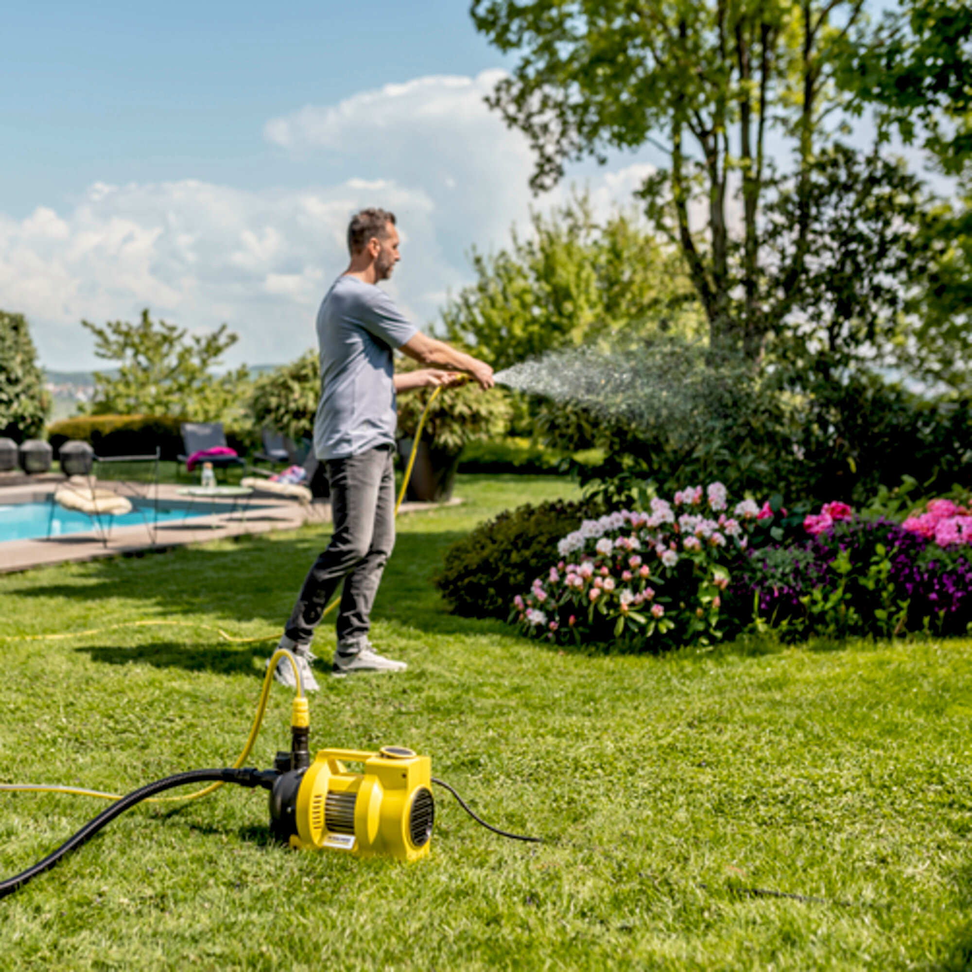 Mann bewässert einen grünen Rasen und Blumen mit einer gelben Wasserpumpe in einem sonnigen Garten.
